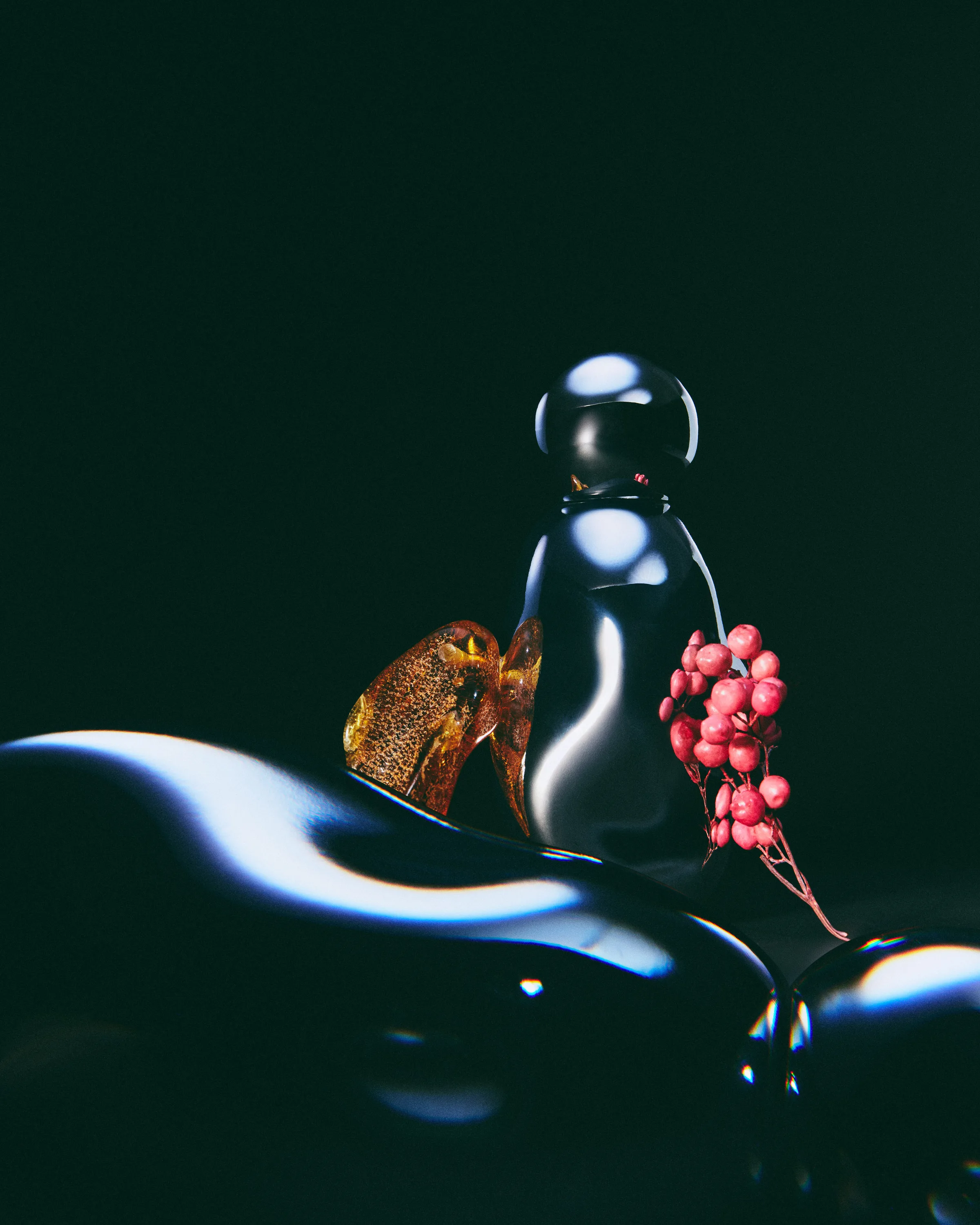 Glossy black perfume bottle with amber resin and pink peppercorns on dark reflective surfaces, for Kylie Cosmetics.