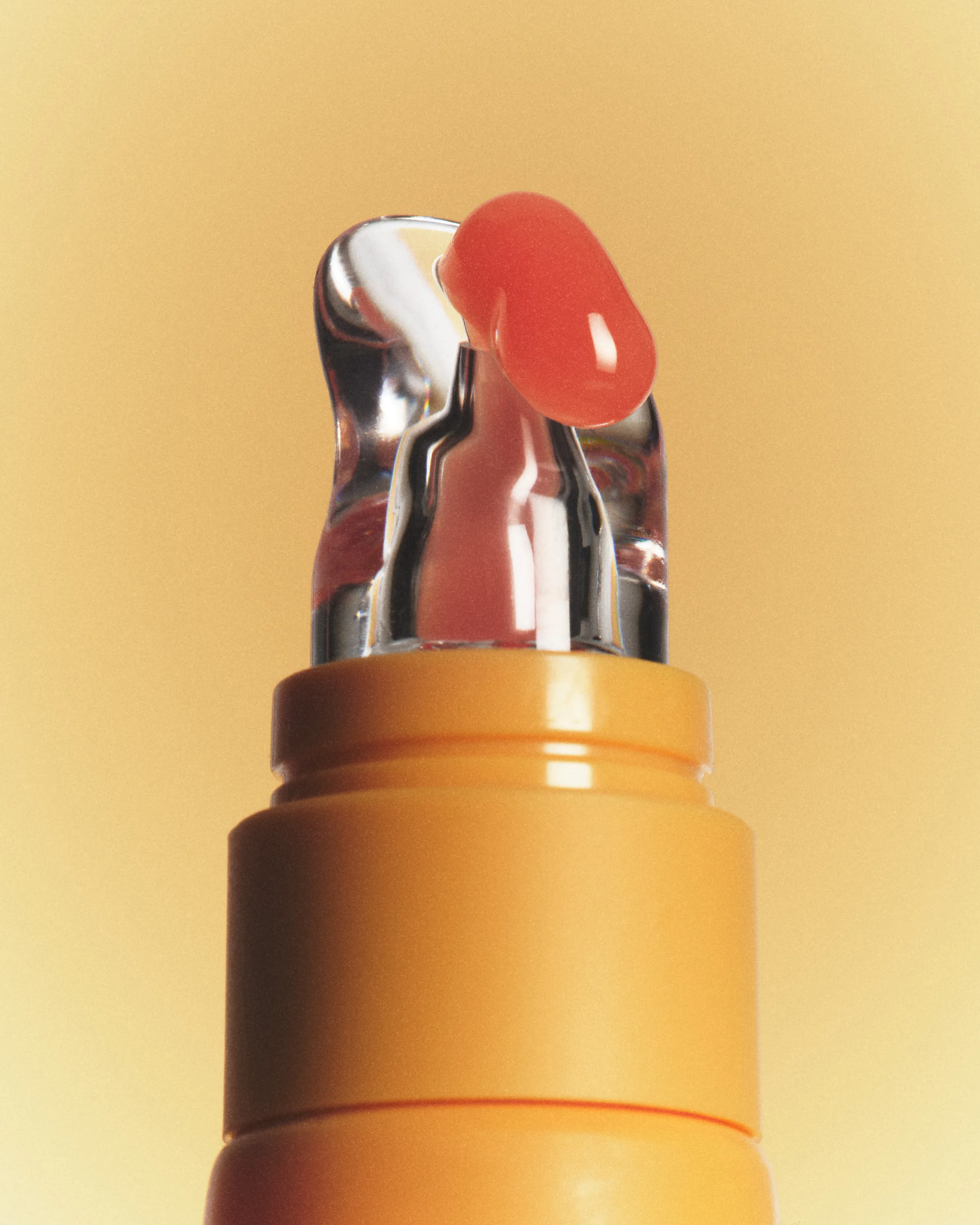 Close-up of an orange lip product tube with a clear applicator tip holding a drop of coral gloss, for Kylie Cosmetics.
