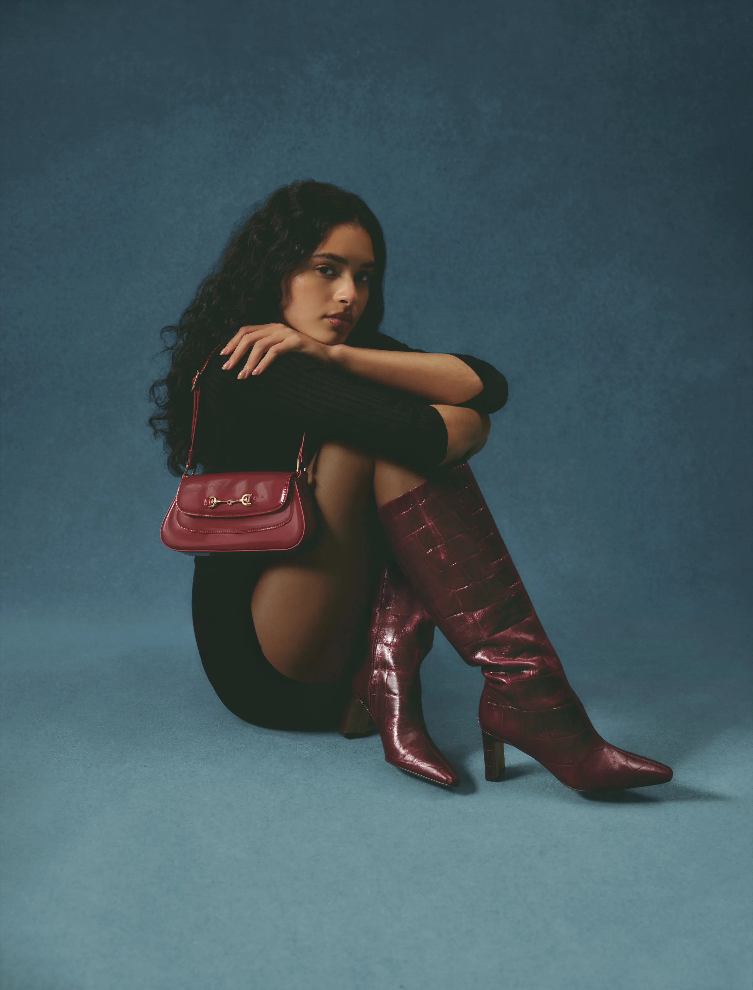 Model seated on a teal backdrop in merlot croc-embossed knee-high boots with a red shoulder bag, for Sam Edelman.