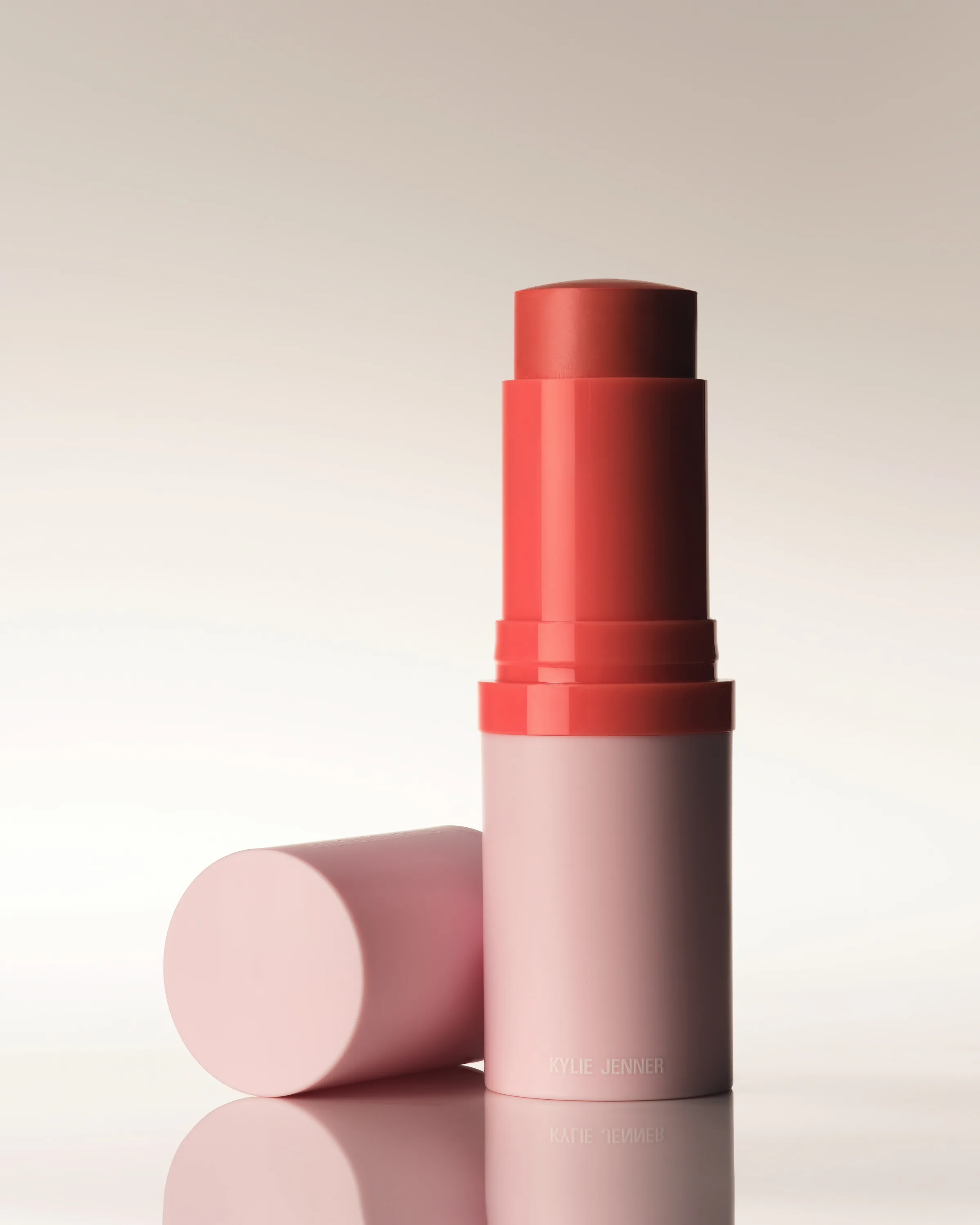 Coral blush stick in a pink tube beside its cap on a reflective surface against a soft grey backdrop, for Kylie Cosmetics.