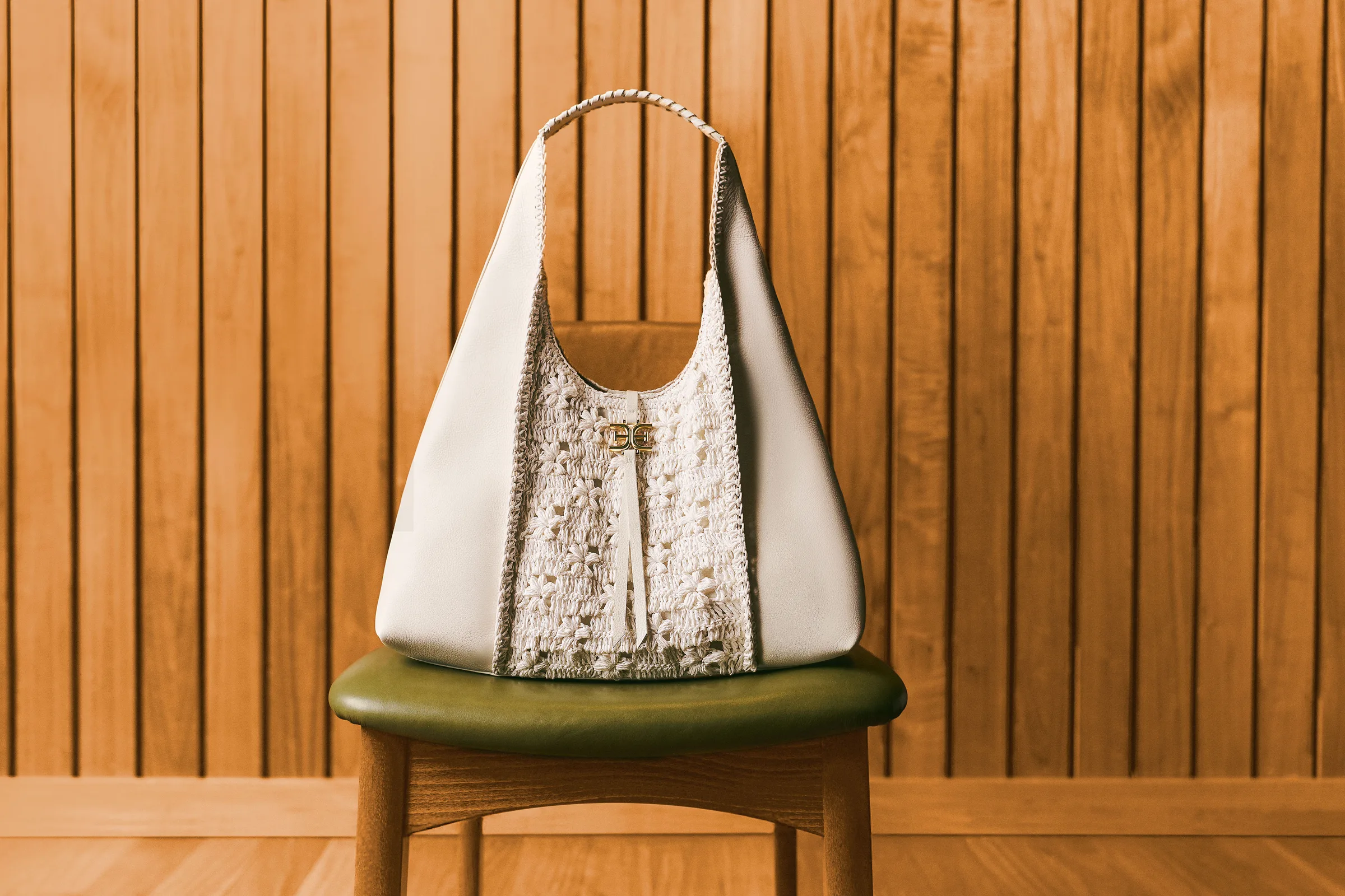 Cream leather hobo bag with crochet panel and gold logo on a green chair against wood paneling, for Sam Edelman.