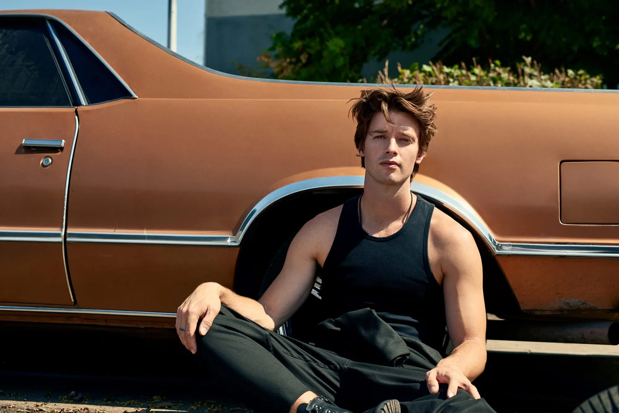 Man in black tank top and trousers sitting against a copper vintage car in sunlight, for Man About Town.