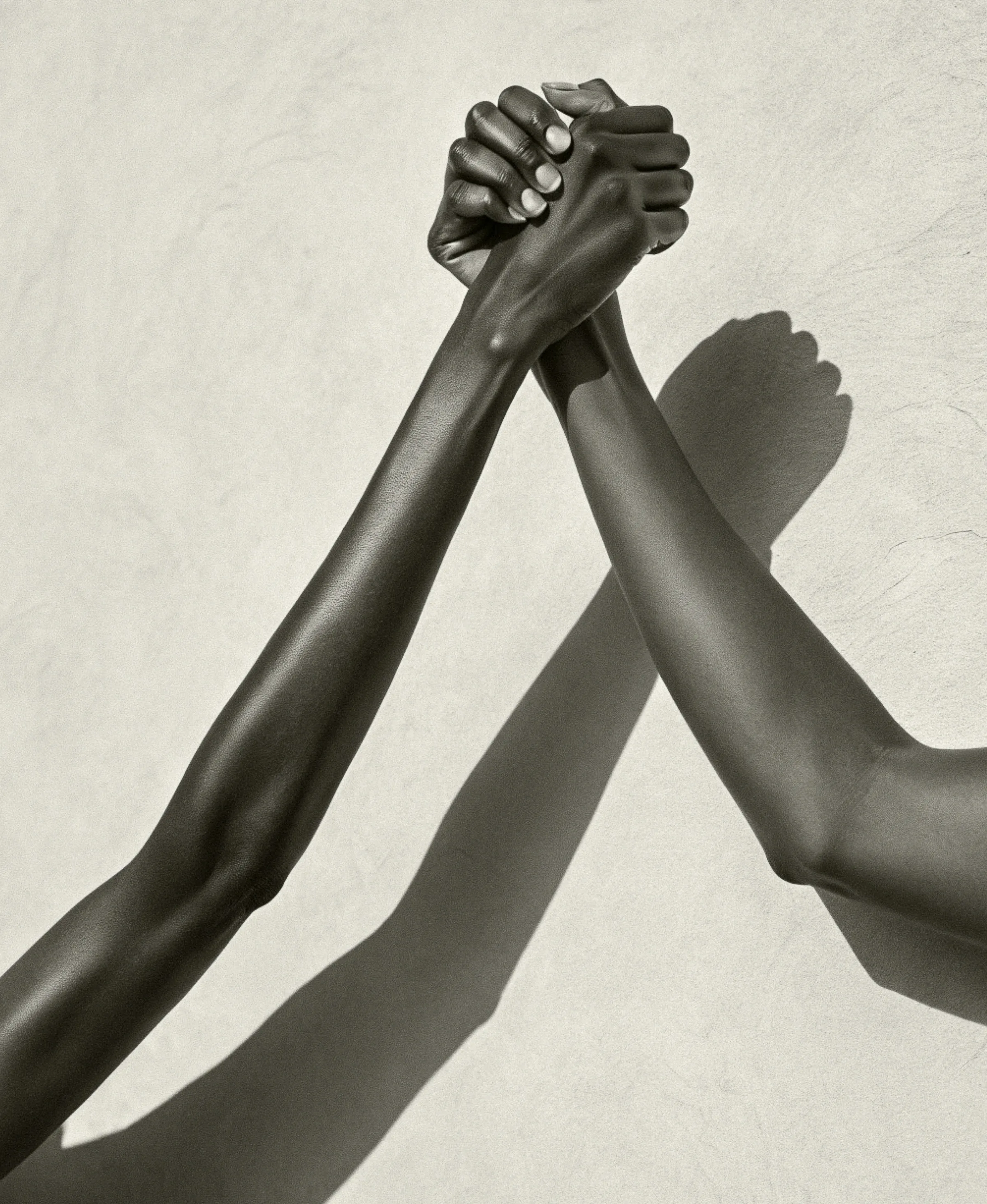 Black-and-white image of two raised arms clasping hands against a textured wall, casting a long shadow. Aesop AI Concept.