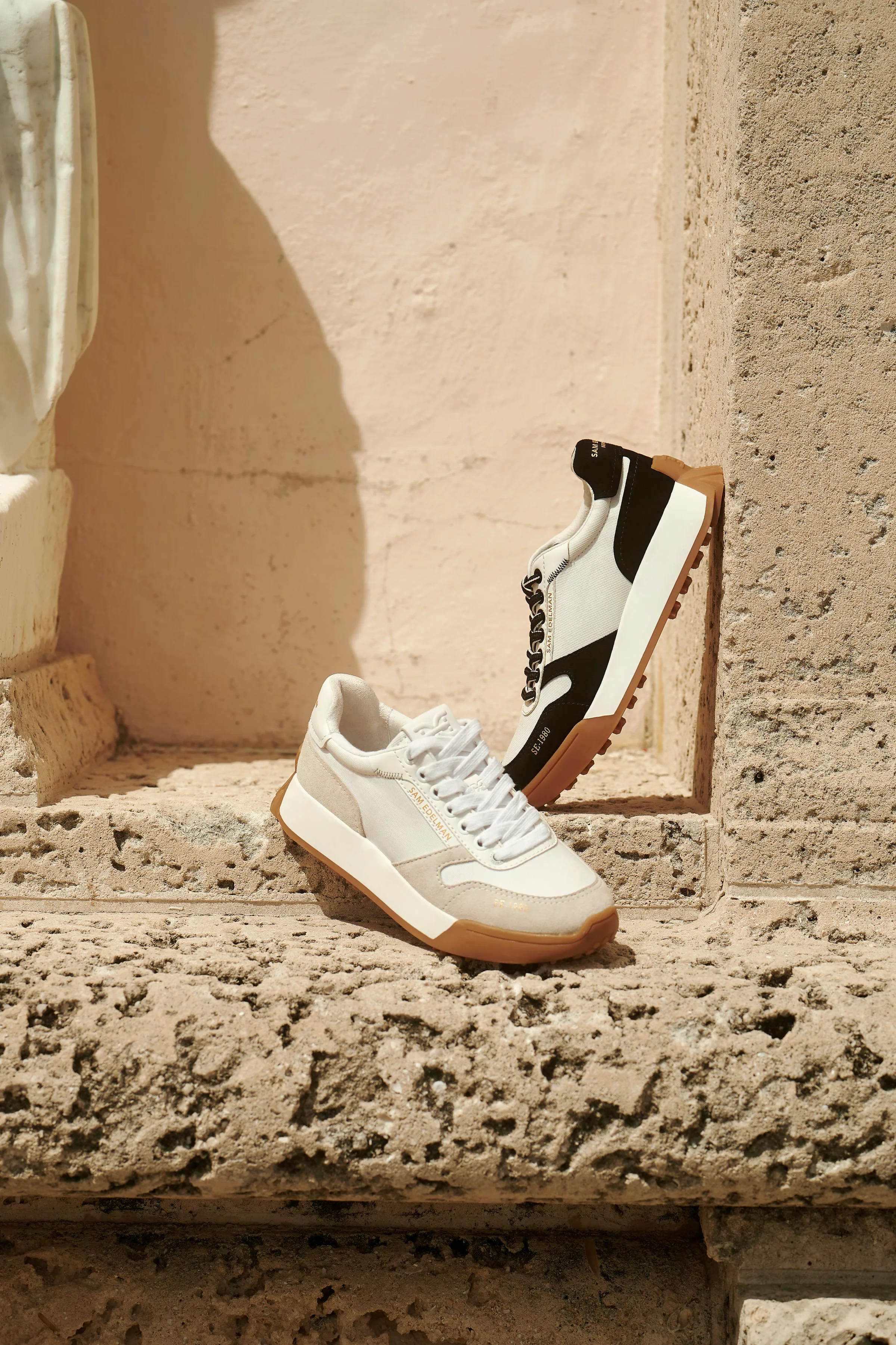 Two retro sneakers, one cream and one black and white, on a sunlit coral stone ledge with gum soles, for Sam Edelman.