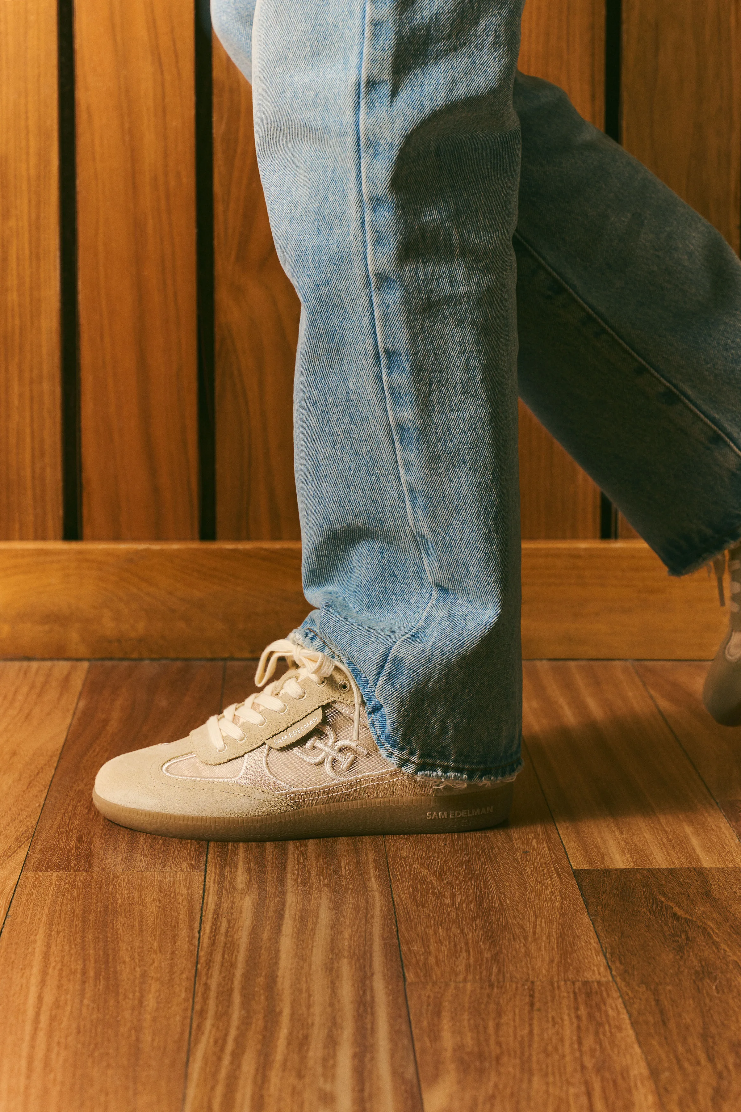 Beige suede lace-up sneakers worn with light wash jeans, mid-stride on a wood floor against slatted wood panels, for Sam Edelman.