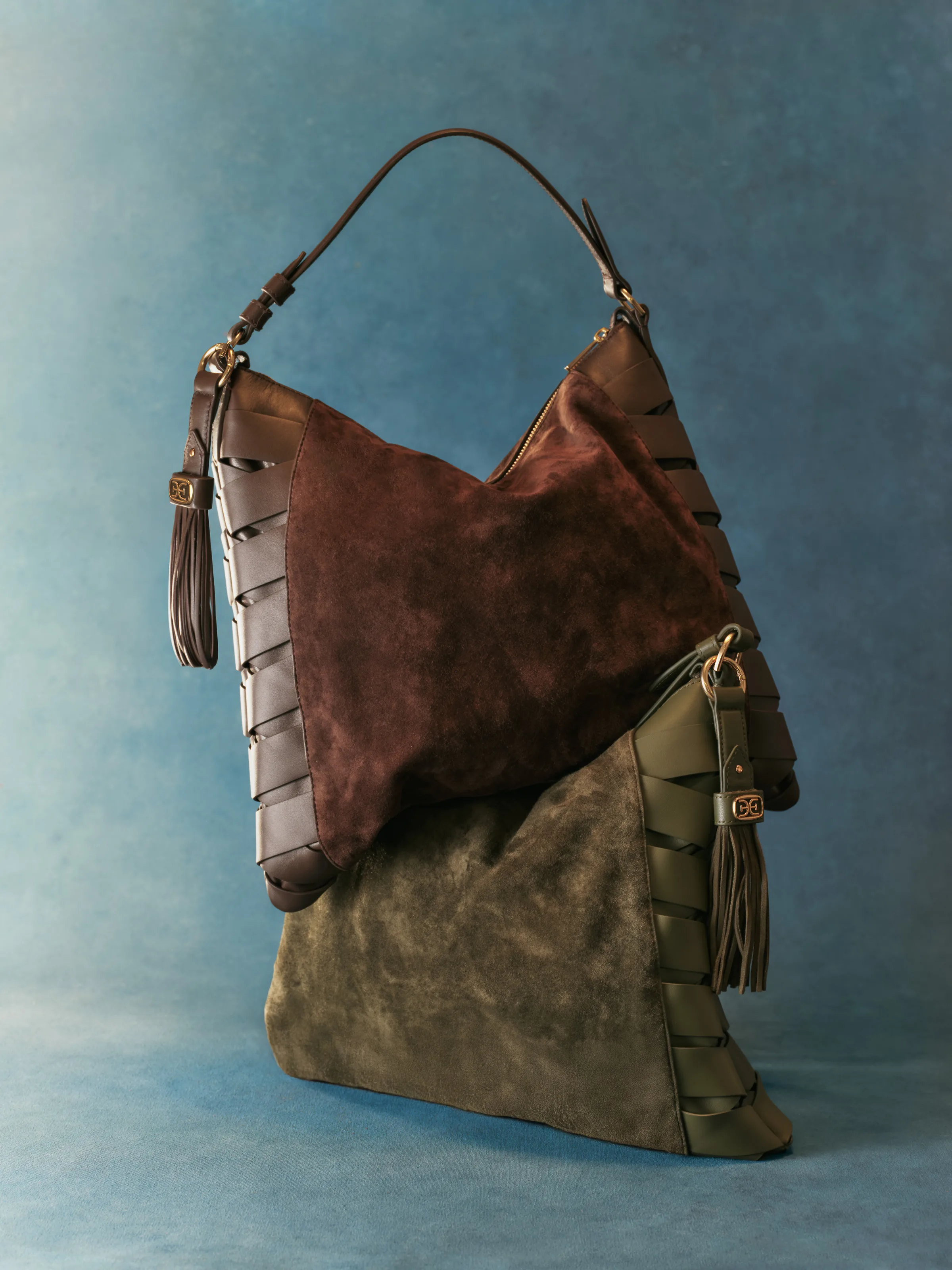 Brown and olive suede hobo bags with woven leather sides and tassels stacked on a blue backdrop, for Sam Edelman.
