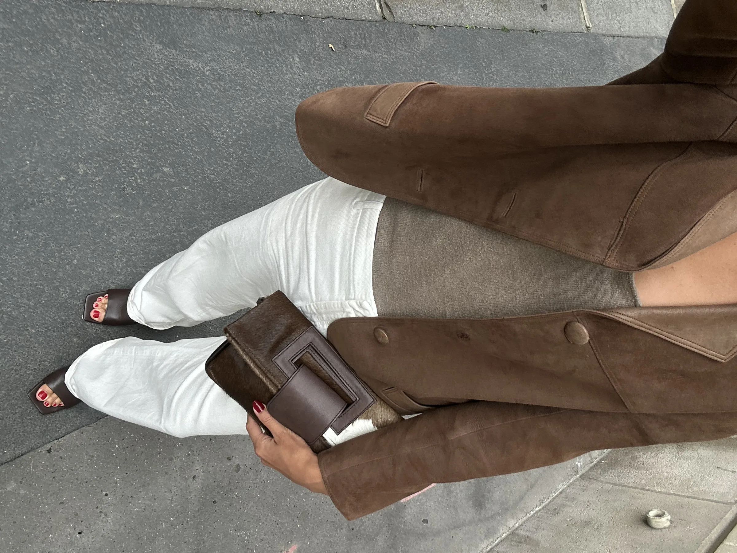Phone-style outfit shot of a brown suede jacket, white jeans and brown pony-hair clutch on a city sidewalk.