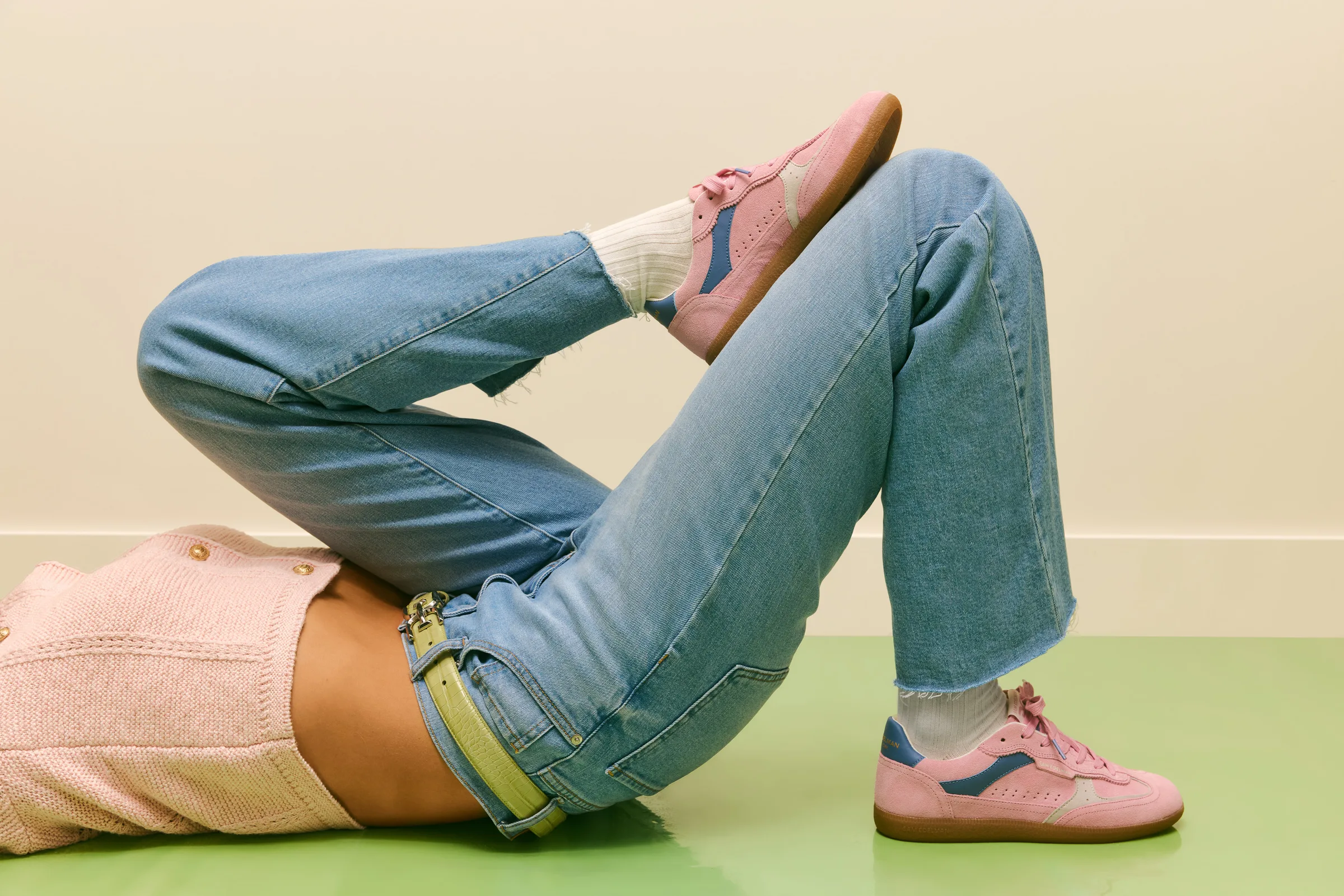 Model lying on a green floor in light-wash jeans, pink knit top and pink suede Kallen sneakers, for Sam Edelman.