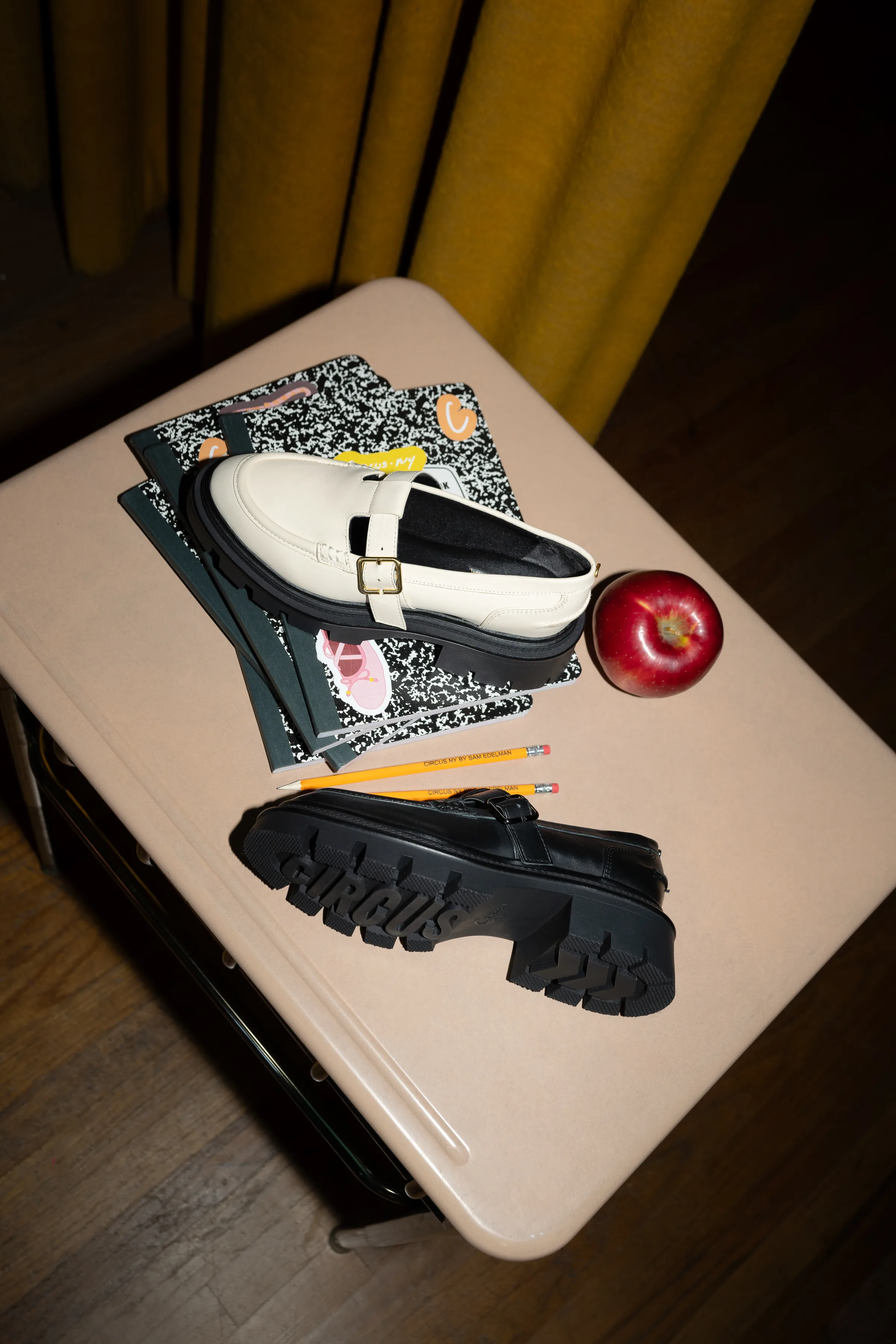 Cream and black lug-sole Mary Jane loafers on a school desk with notebooks, pencils and a red apple, for Sam Edelman.