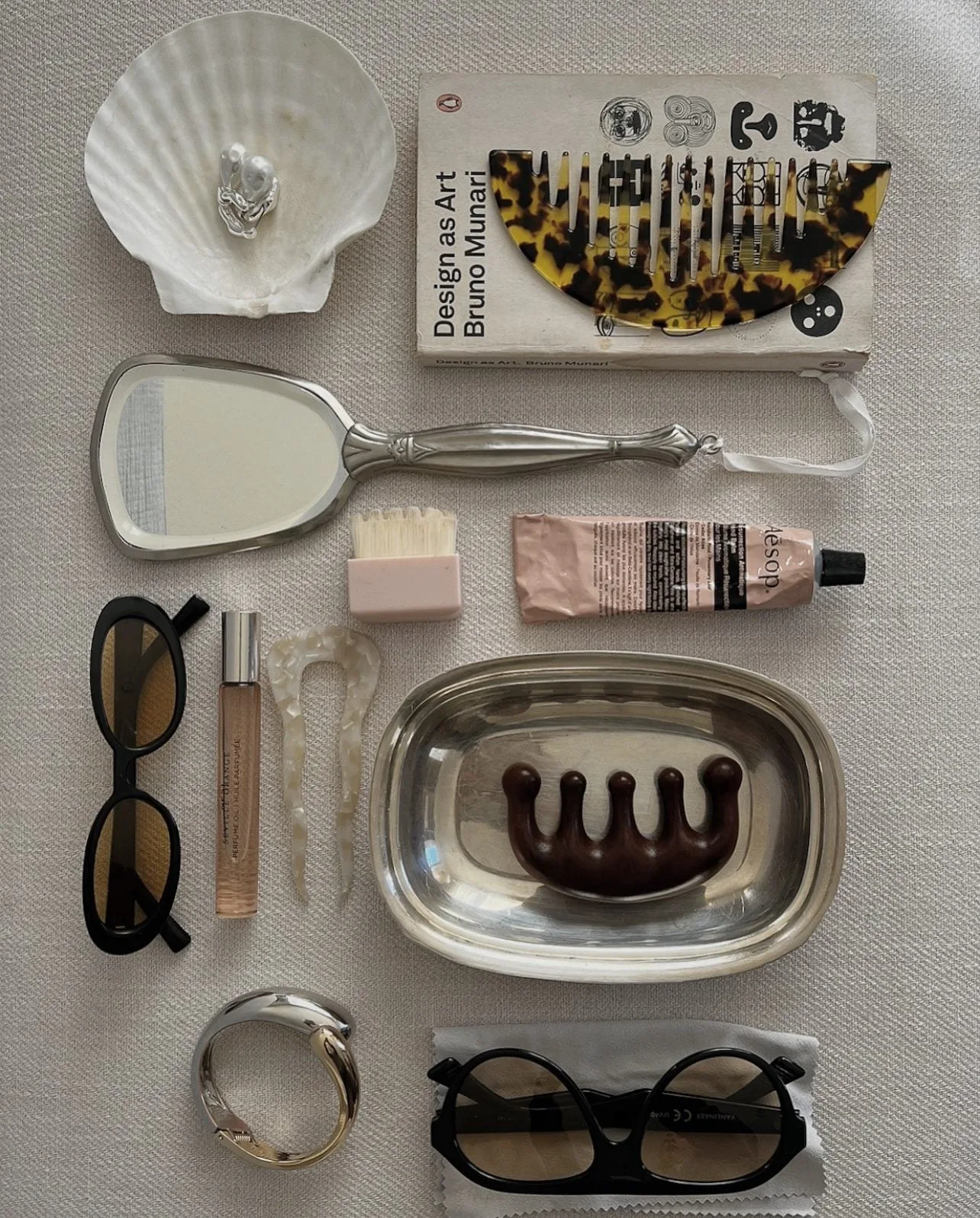 Flat lay of a silver hand mirror, tortoiseshell comb, sunglasses, perfume, Aesop tube and silver cuff on linen.