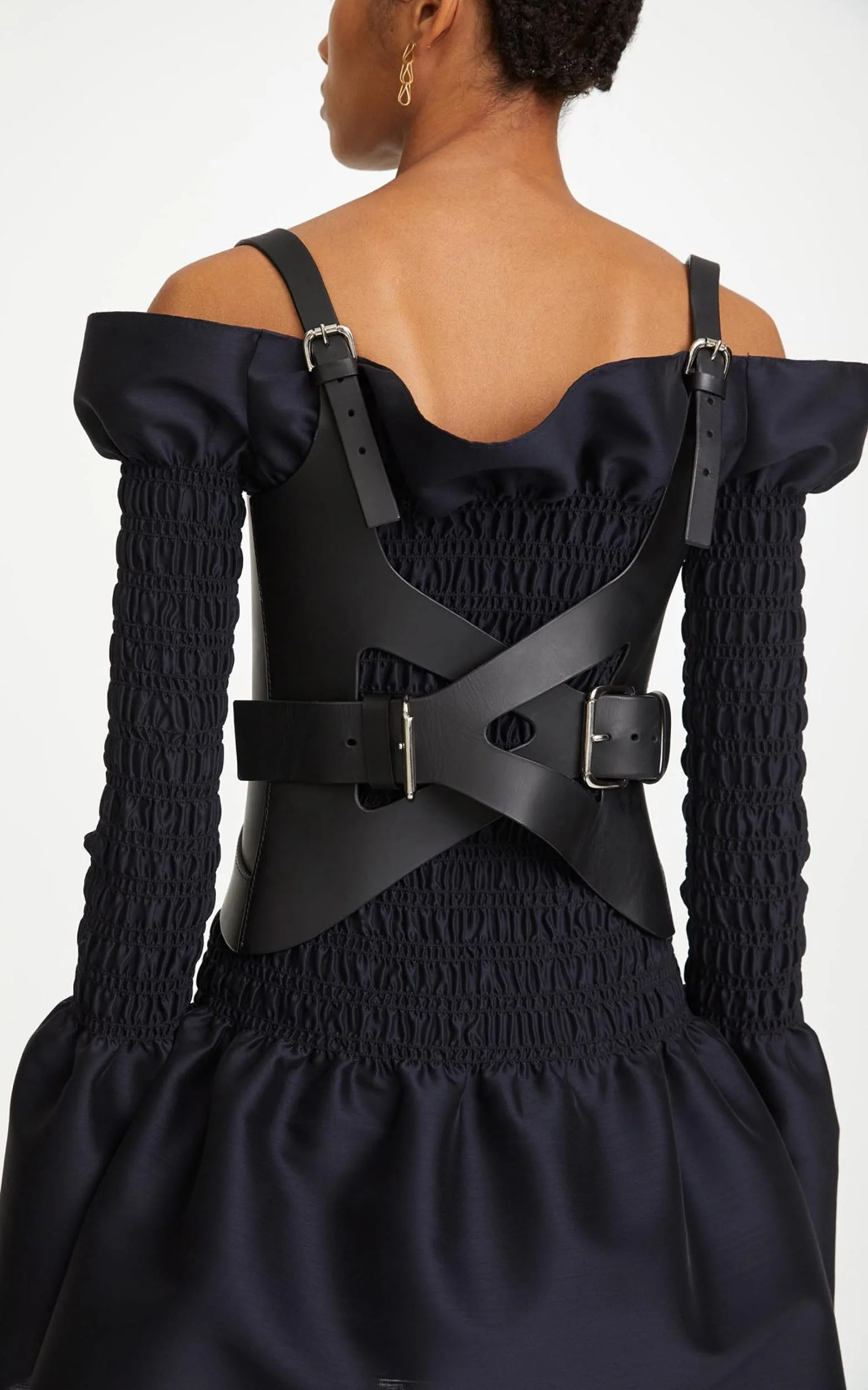 Back view of a model in a navy off-shoulder smocked dress with a black leather buckled harness, for Dion Lee.