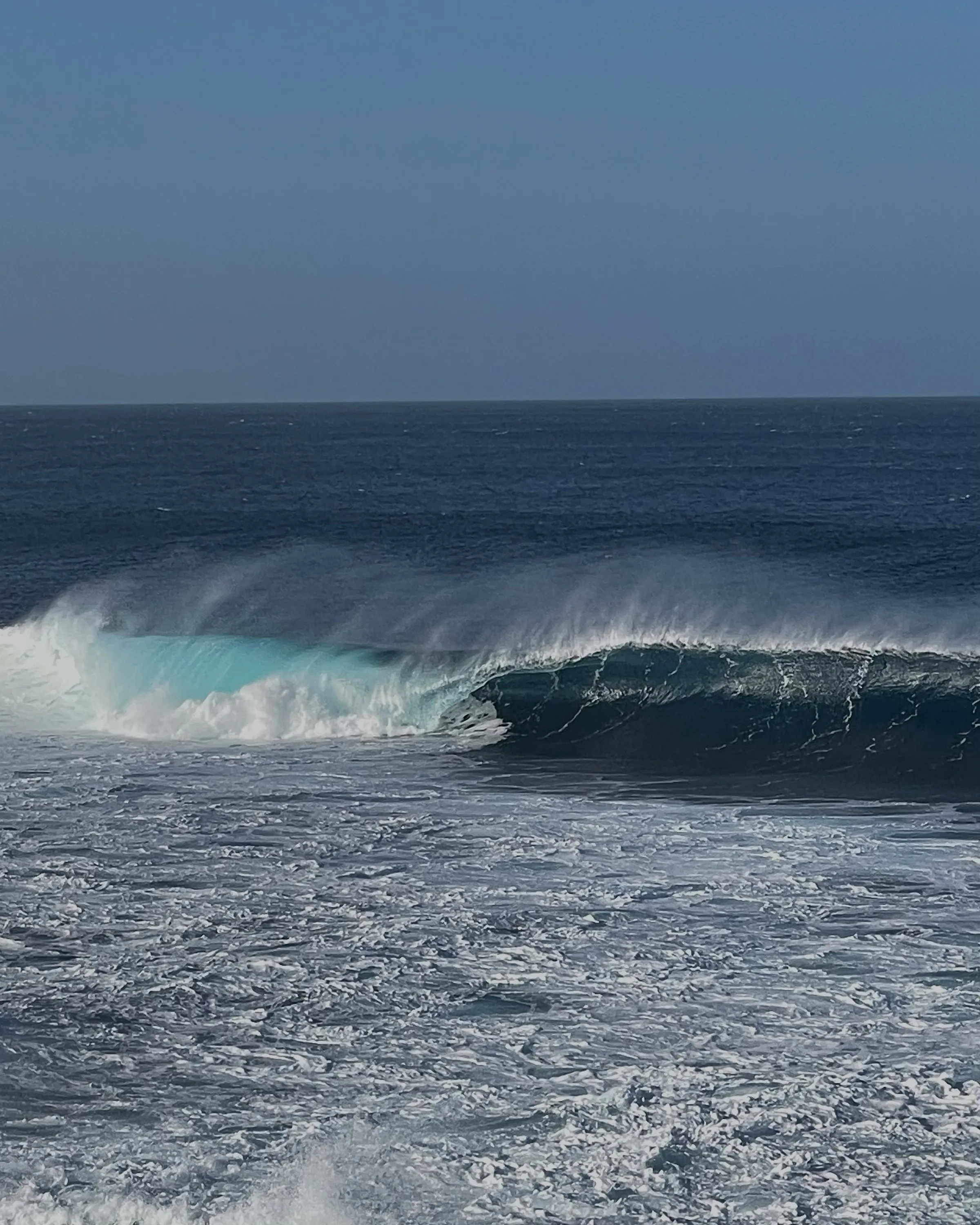 Turquoise ocean wave curling and breaking under a clear sky, with white foam in the foreground.