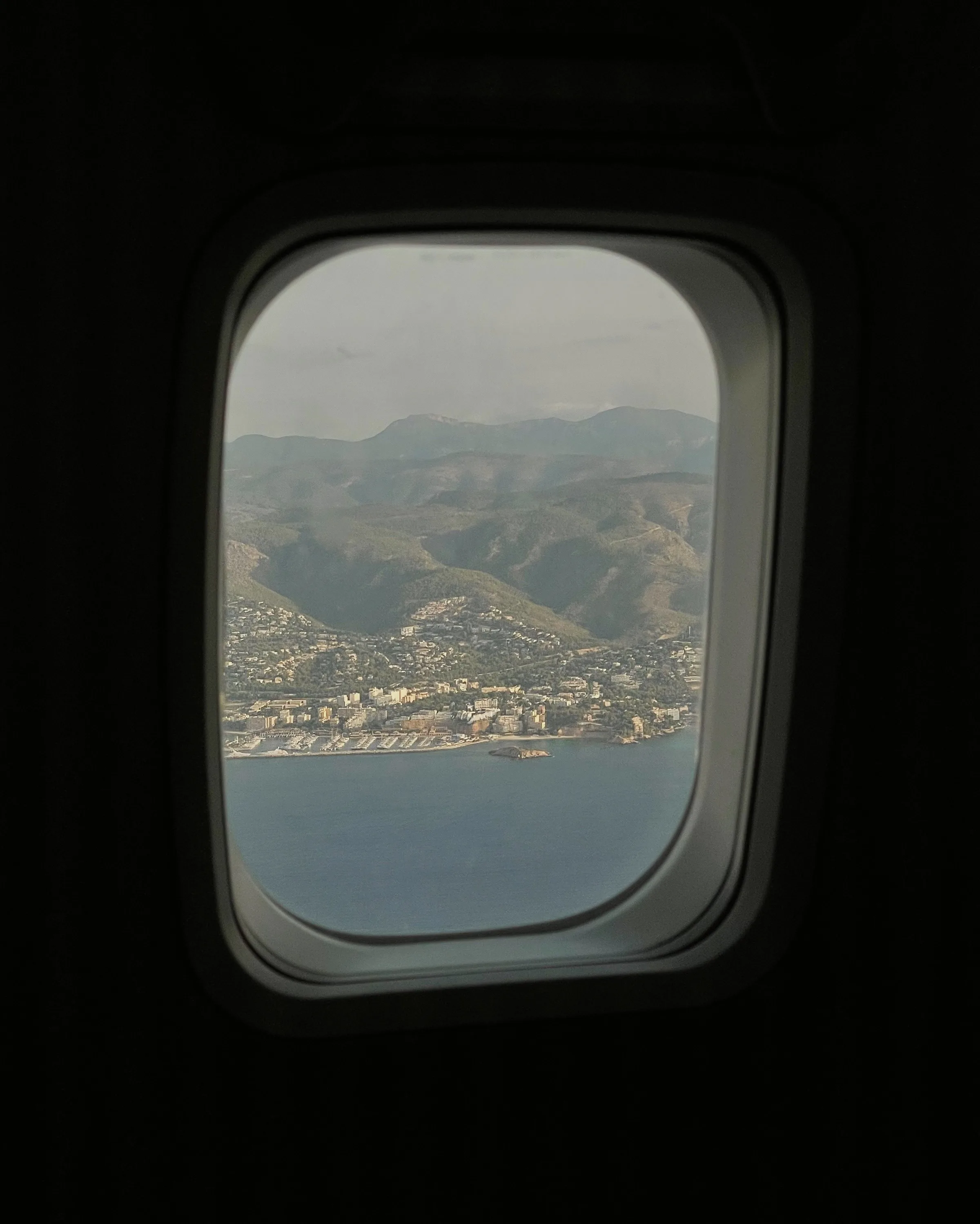 View through an airplane window of a coastal town, mountains and blue sea below.