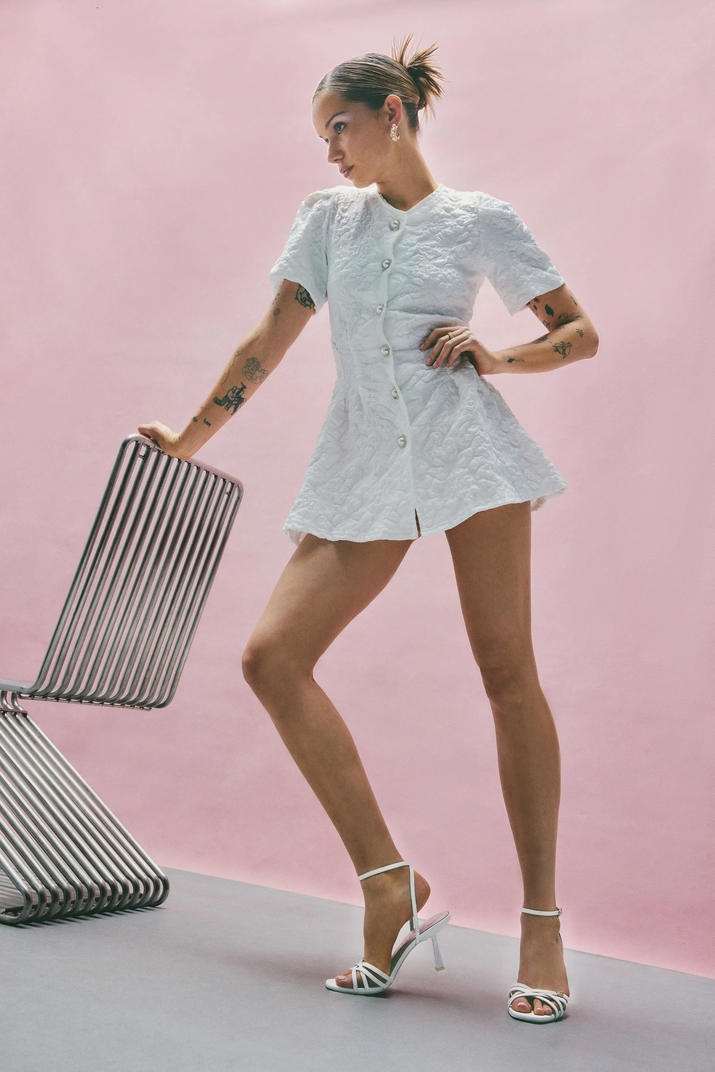 Model in a white quilted mini dress and white strappy heeled sandals beside a metal chair on pink backdrop, for Sam Edelman.