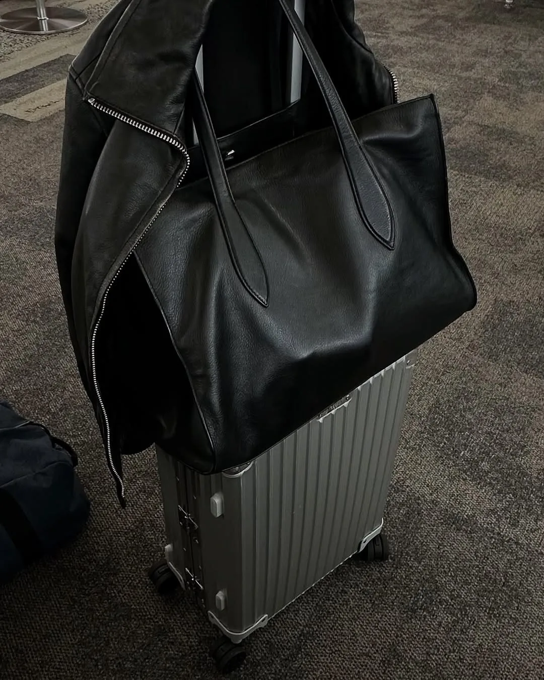 Black leather tote and leather jacket draped over a silver ribbed suitcase on airport carpet.