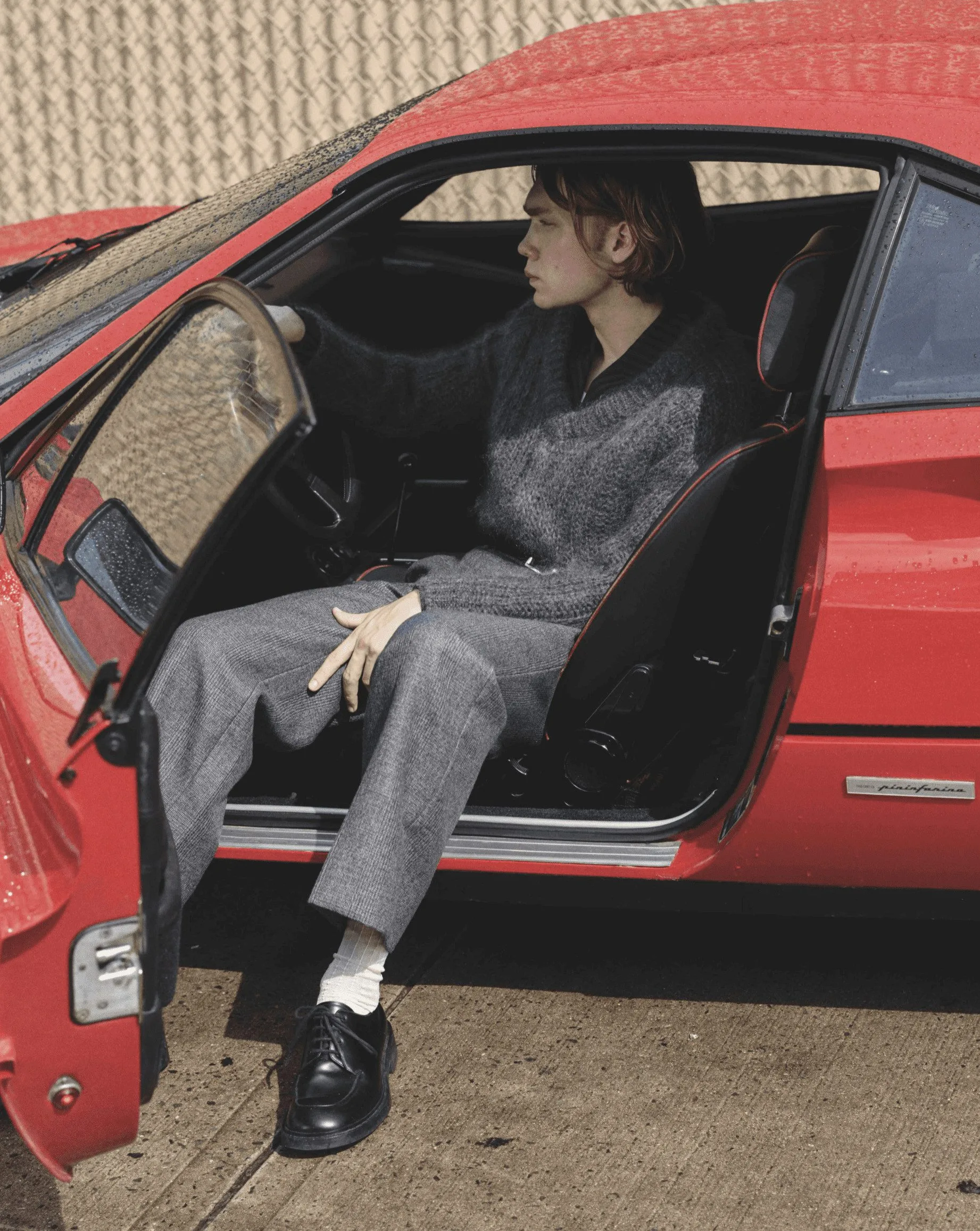 Young man in grey mohair sweater and tweed trousers sitting in a red sports car with the door open, for L'Officiel Hommes.