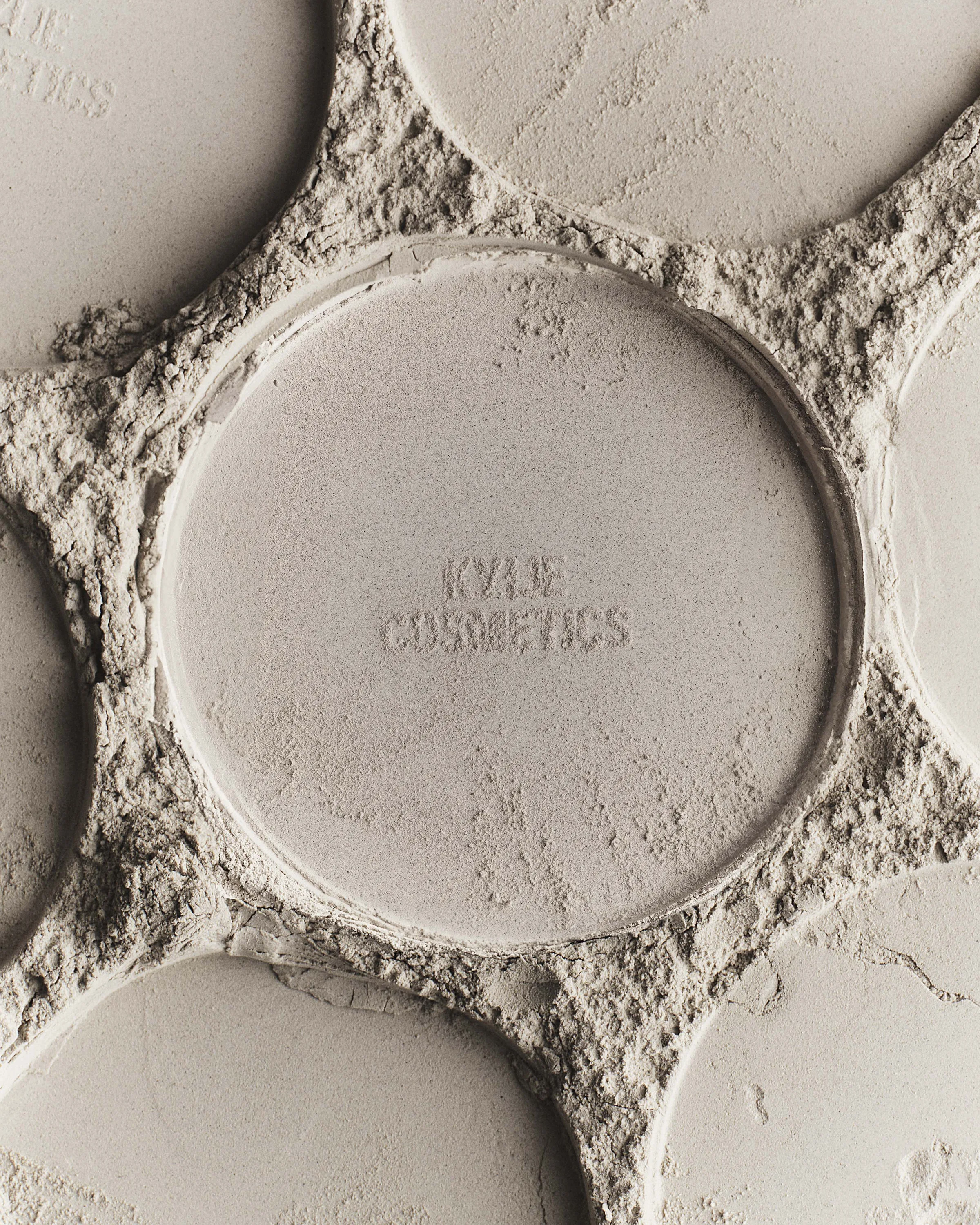 Close-up of beige pressed powder pans embossed with the Kylie Cosmetics name, surrounded by crumbled powder, for Kylie Cosmetics.