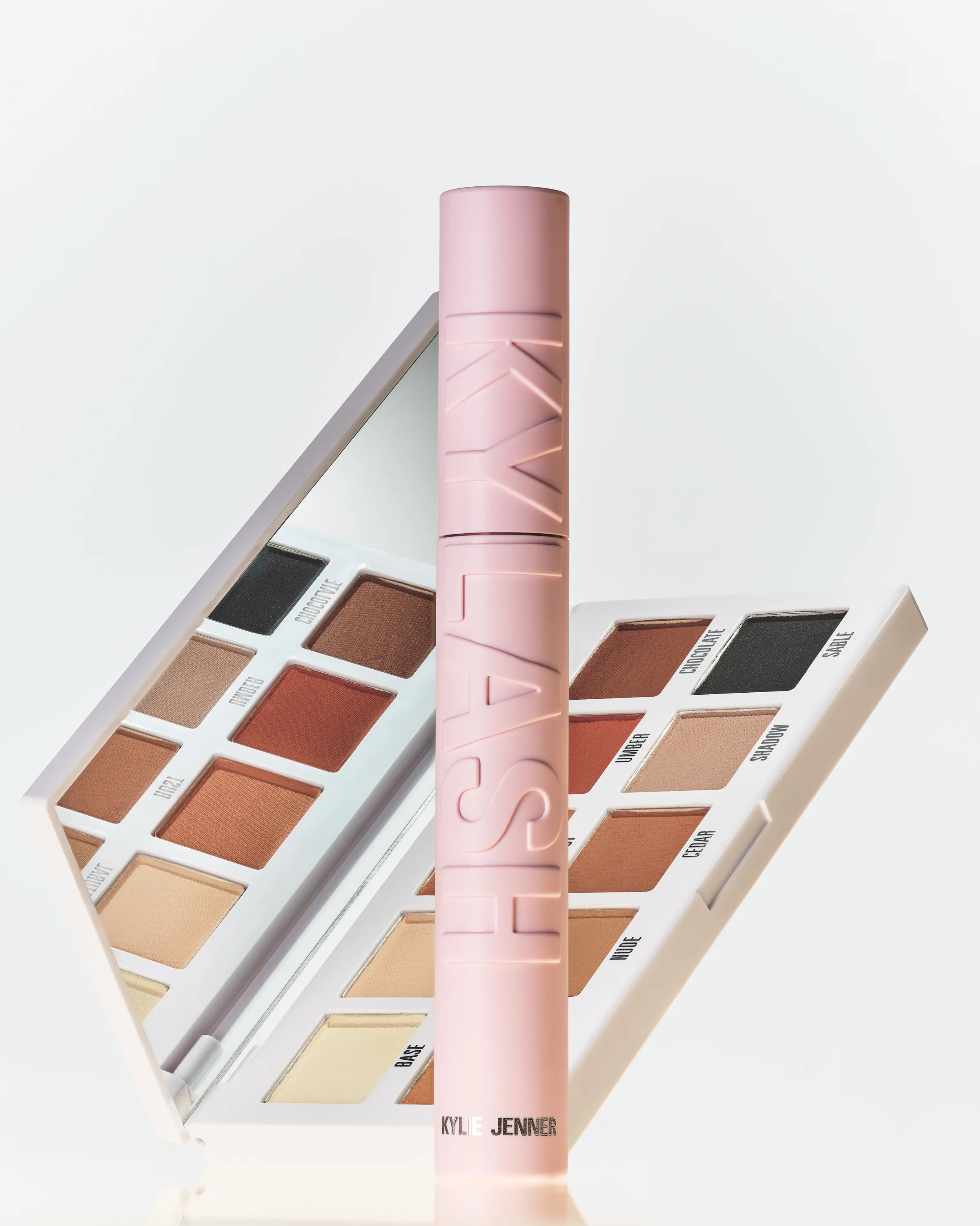 Pink Kylash mascara tube standing before an open brown matte eyeshadow palette on white, for Kylie Cosmetics.