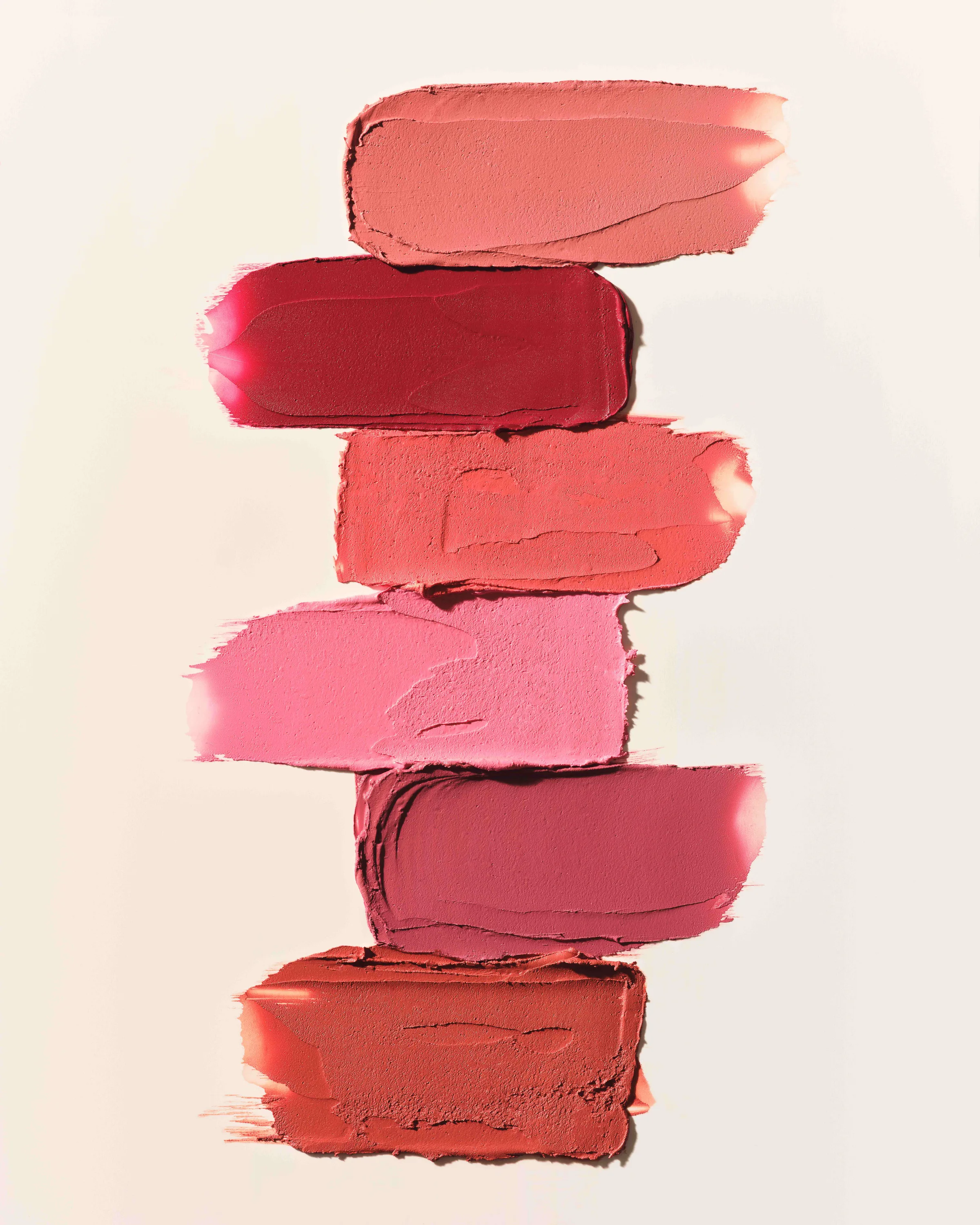 Six lipstick swatches in coral, red, pink and berry shades smeared in a stack on a cream background, for Kylie Cosmetics.