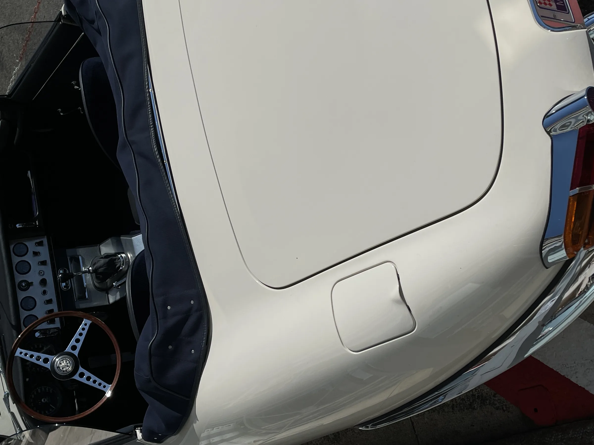 Cream vintage convertible seen from above the rear deck, with navy soft top folded and a wood-rimmed steering wheel.