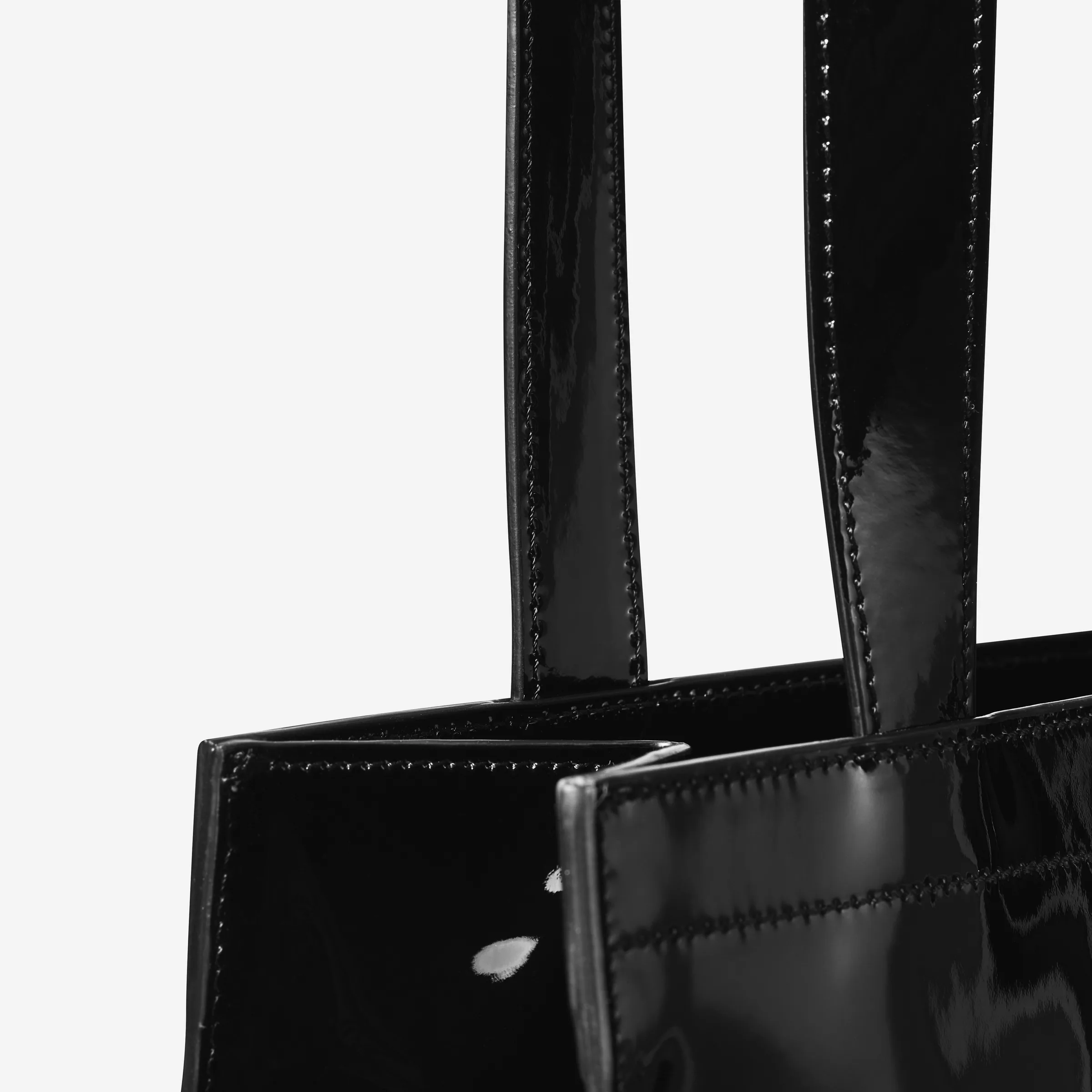 Close-up of a black patent leather tote's stitched handles and top edge on a white background, for Strata.
