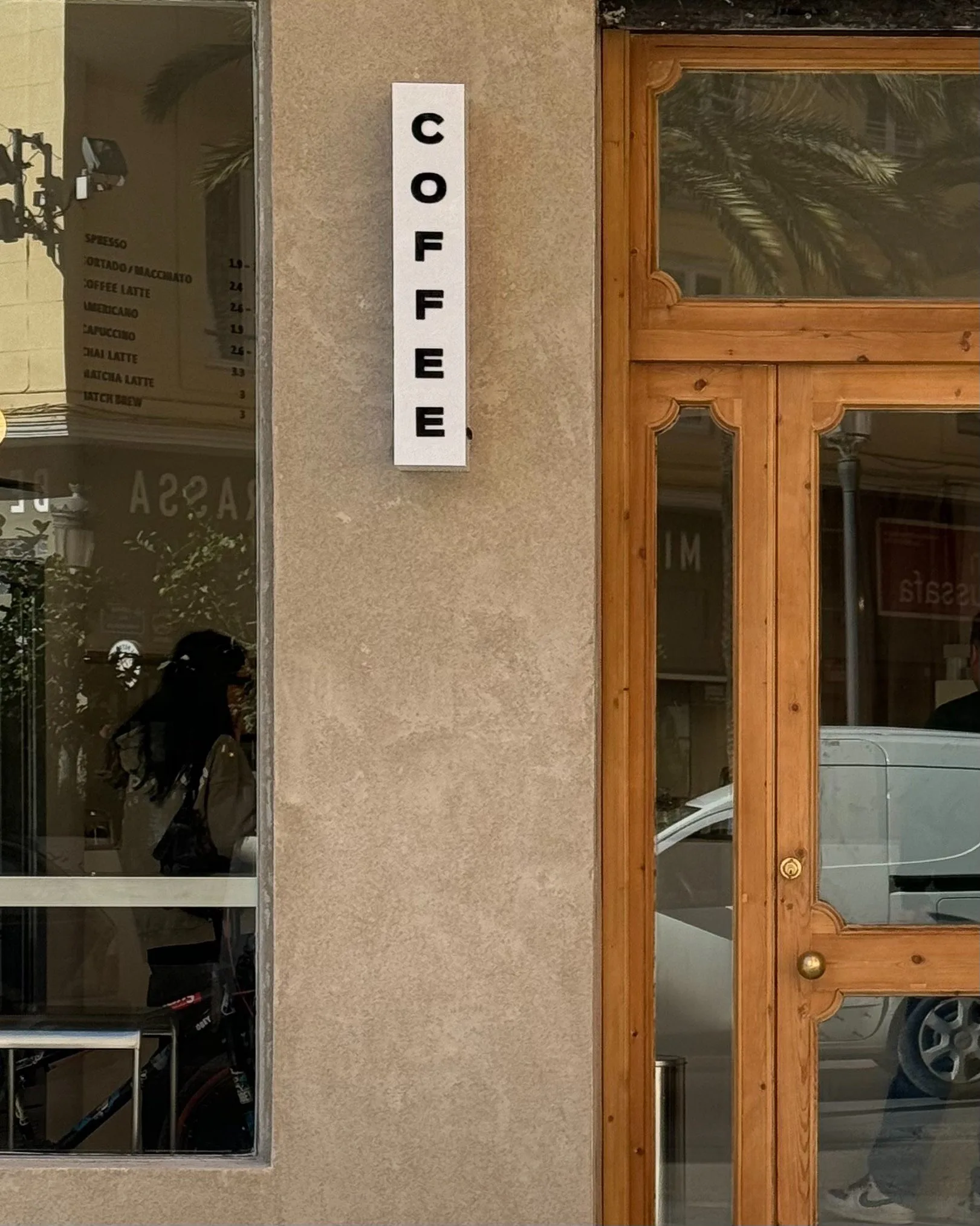 Vertical white COFFEE sign on a stucco wall beside a wooden glass door and a menu window.
