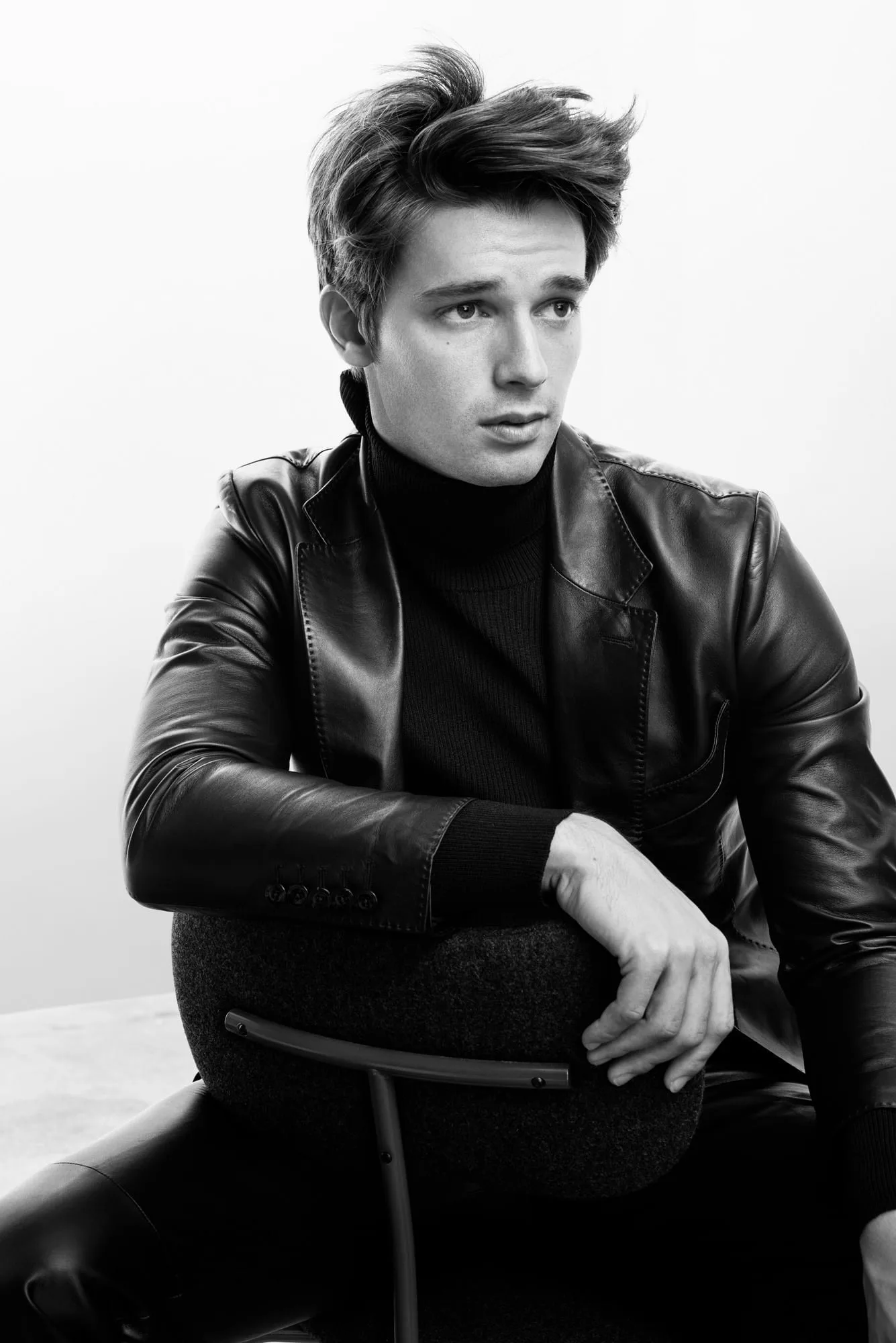 Black and white portrait of a man in a leather blazer and turtleneck leaning on a chair back, for Man About Town.