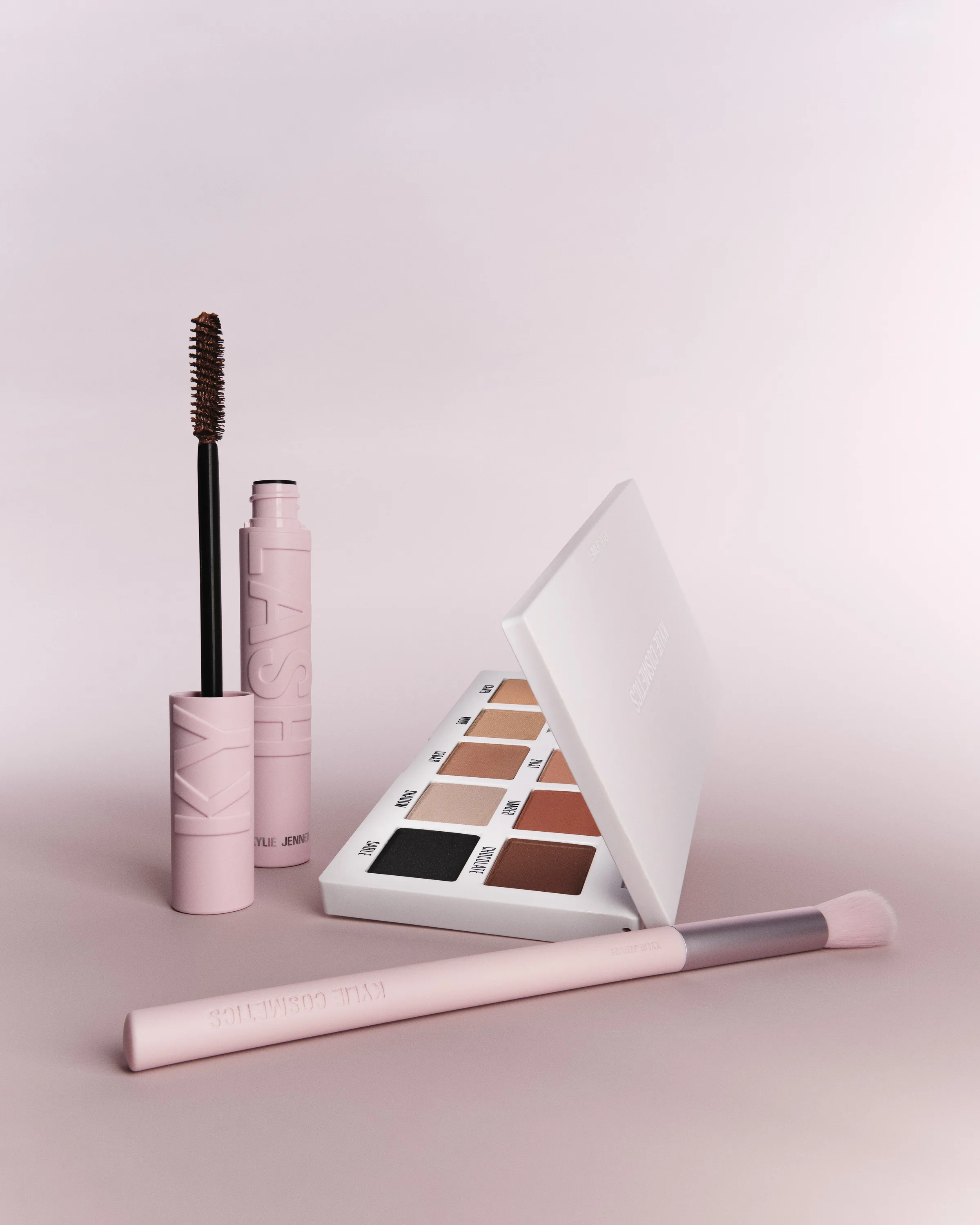 Pink mascara, open eyeshadow palette and pink makeup brush in a still life on a soft pink backdrop, for Kylie Cosmetics.
