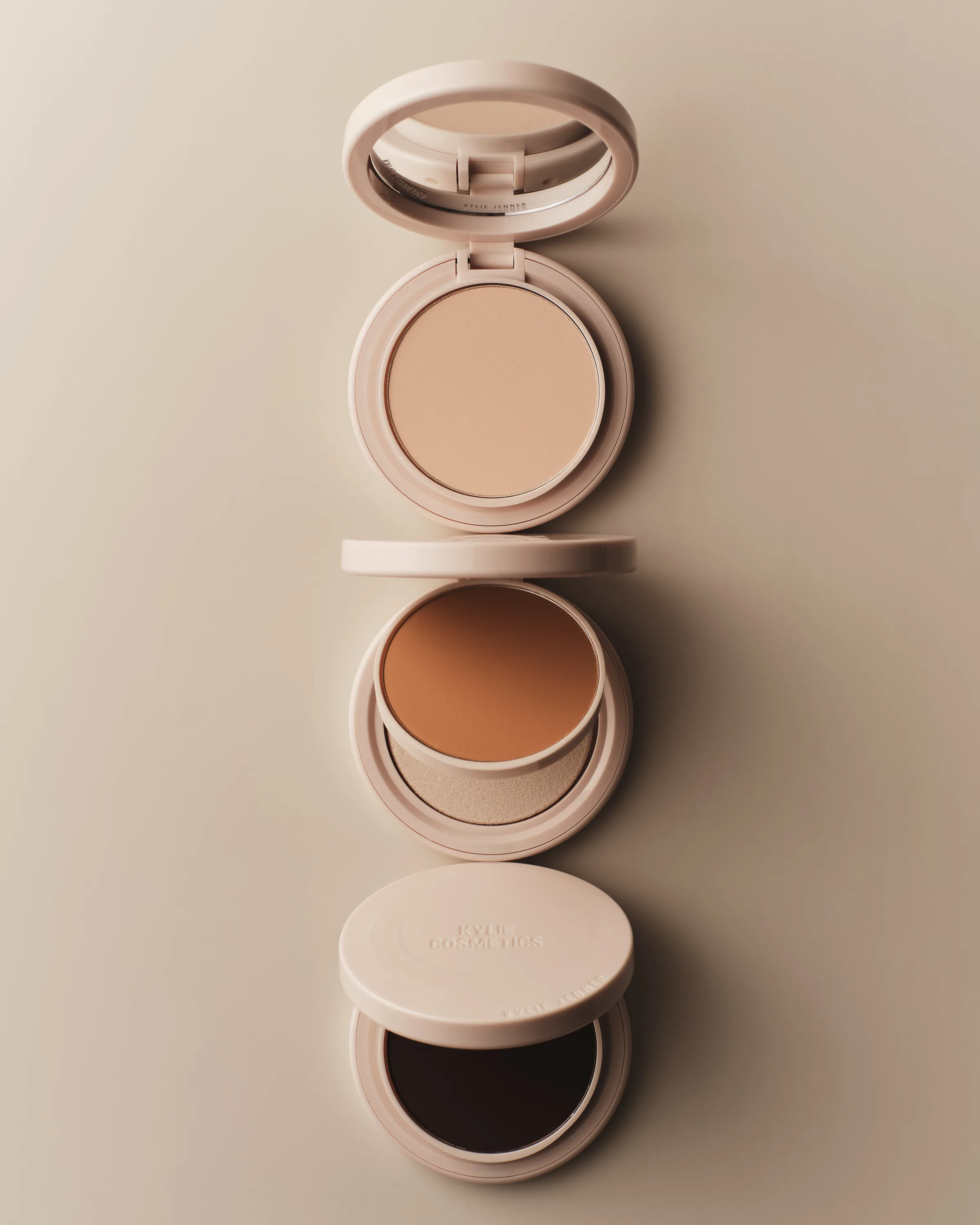Three open beige powder compacts in light, medium and deep shades stacked on a cream backdrop, for Kylie Cosmetics.