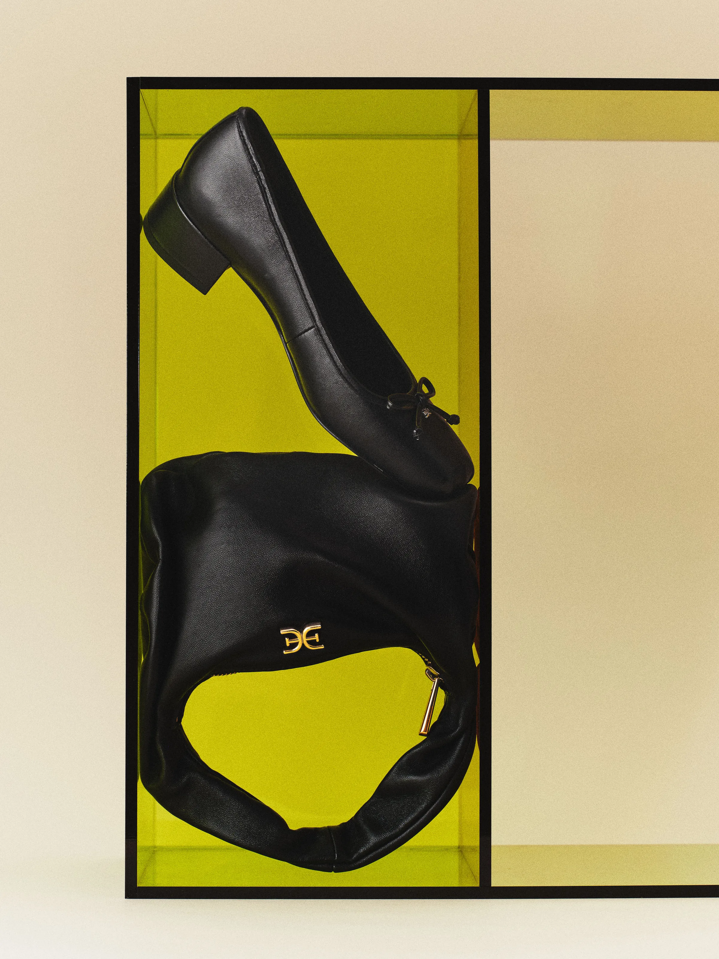 Black leather bow ballet flat resting on a slouchy black bag with gold logo inside a yellow acrylic box, for Sam Edelman.