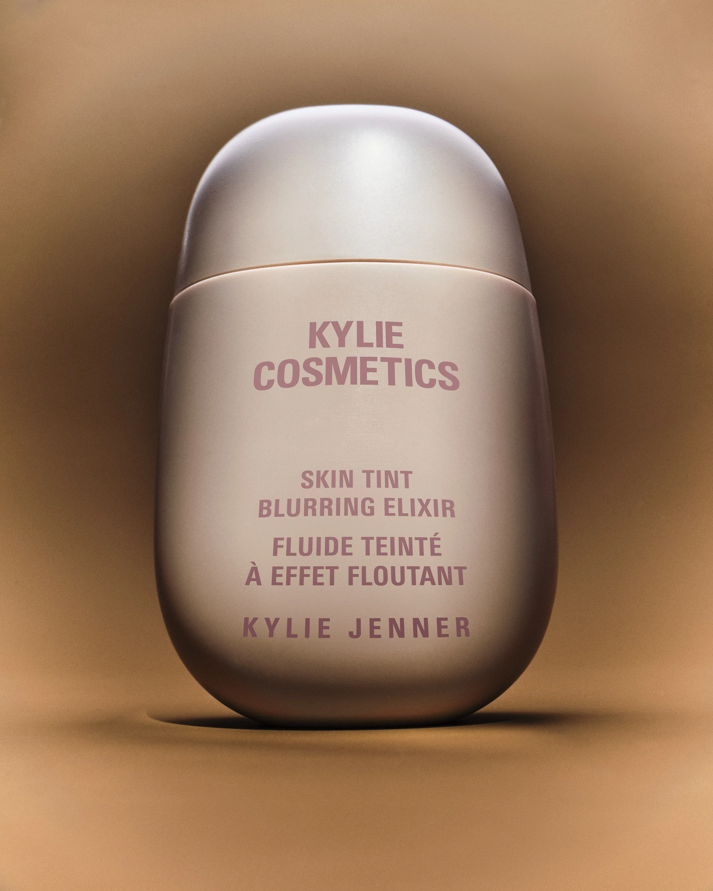 Pale pink Skin Tint Blurring Elixir bottle standing on a warm tan backdrop with soft shadow, for Kylie Cosmetics.