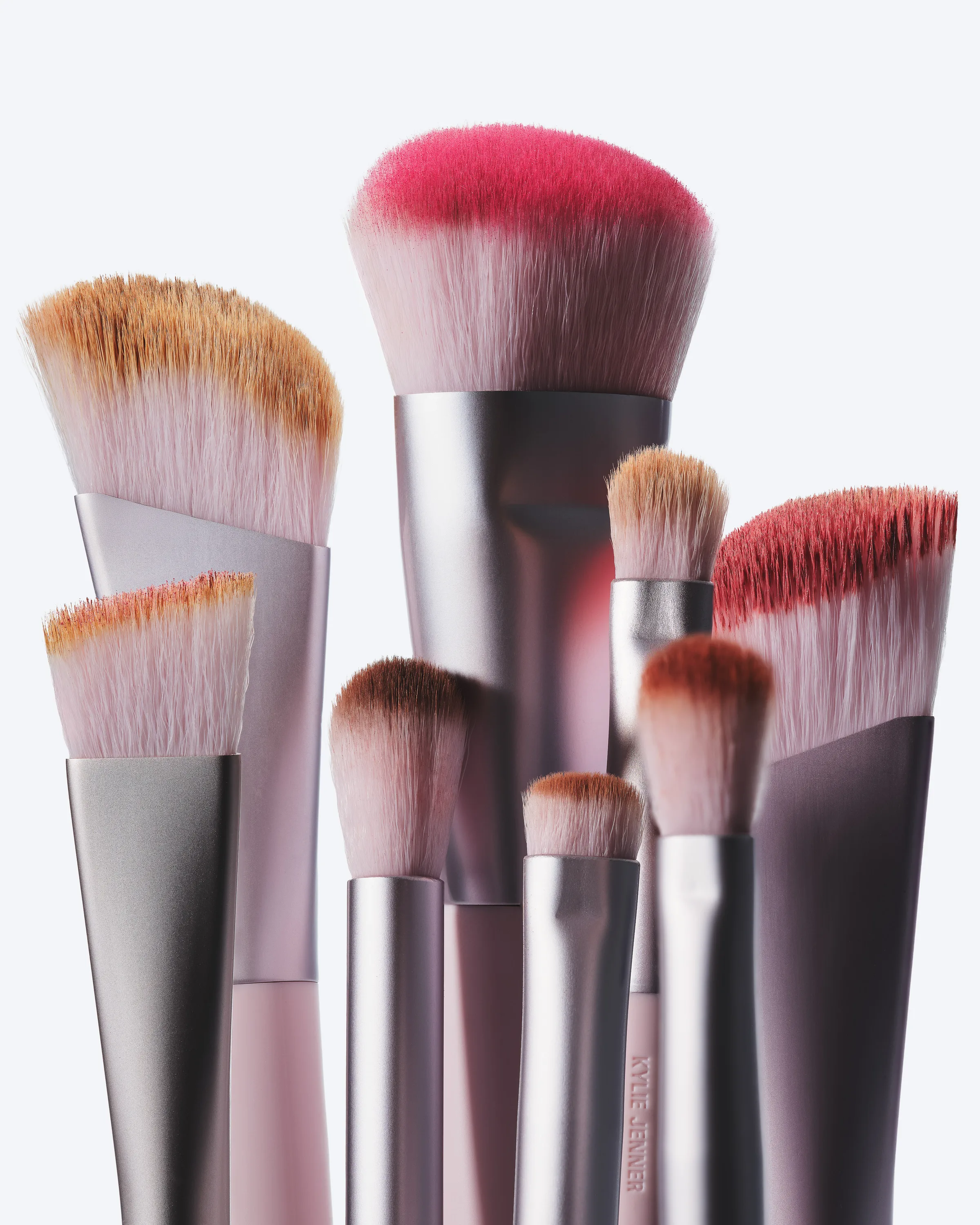 Group of makeup brushes with pink-tipped bristles and metallic mauve handles on a pale grey background, for Kylie Cosmetics.