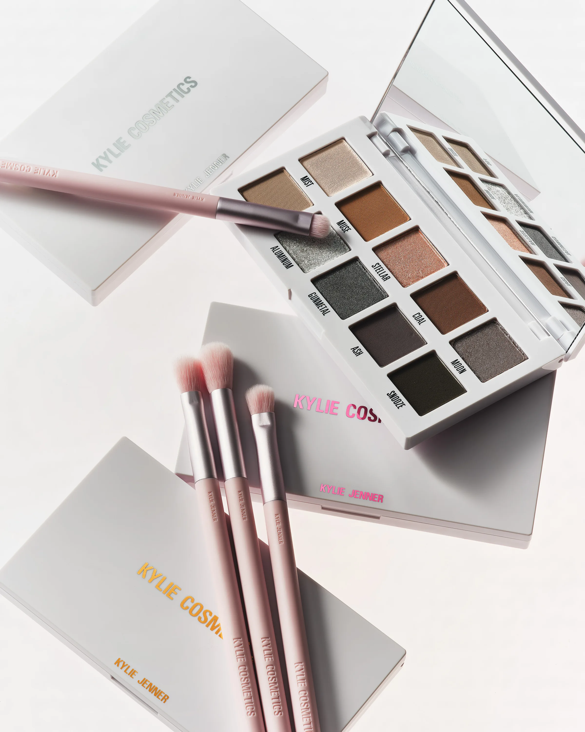 Open neutral eyeshadow palette with mirror, closed white palettes and four pink makeup brushes on white, for Kylie Cosmetics.