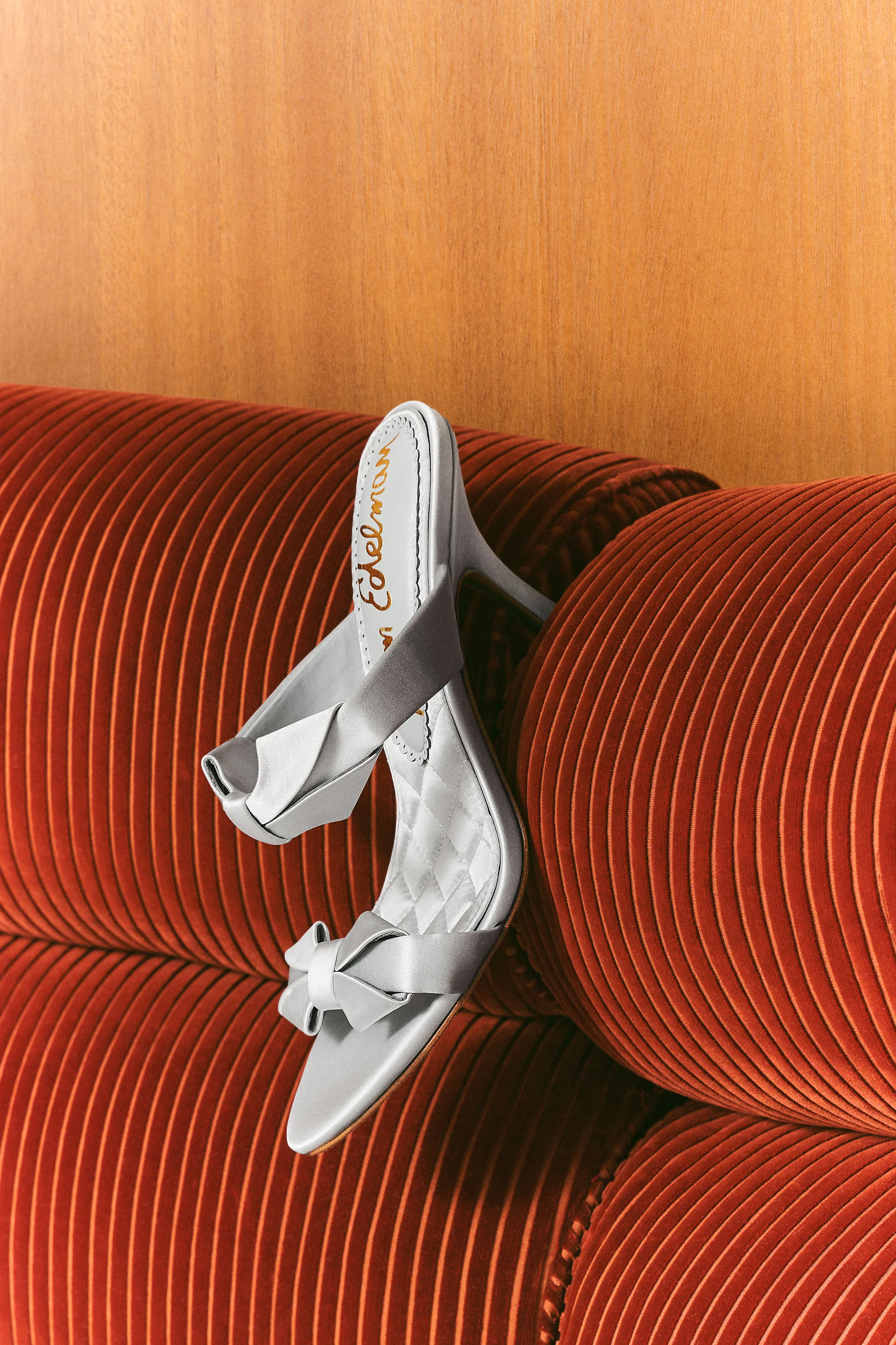 Silver satin bow-detail heeled sandals resting on a rust corduroy sofa against a wood wall, for Sam Edelman.
