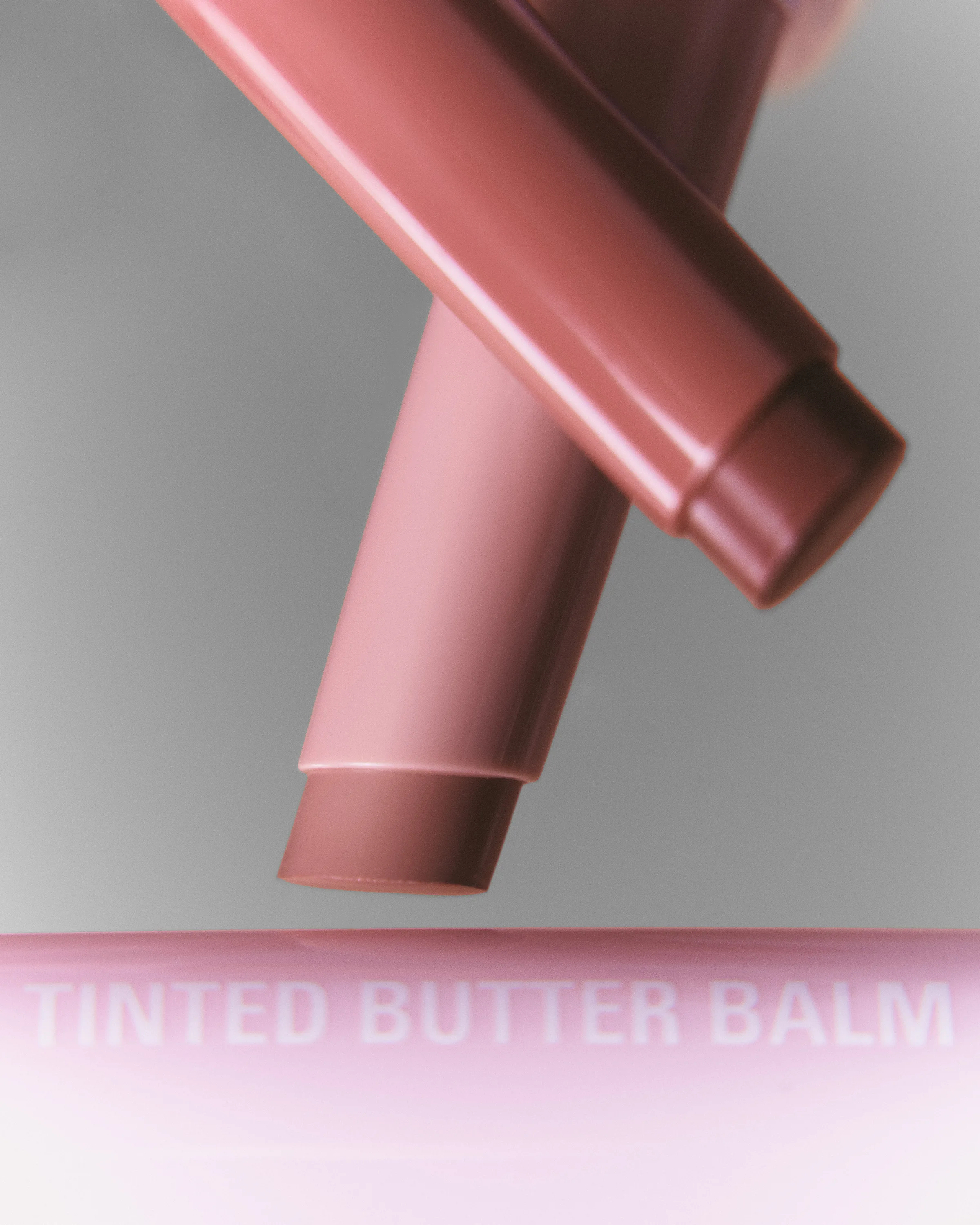 Two pink Tinted Butter Balm lip tubes suspended over a pink surface against a grey background, for Kylie Cosmetics.