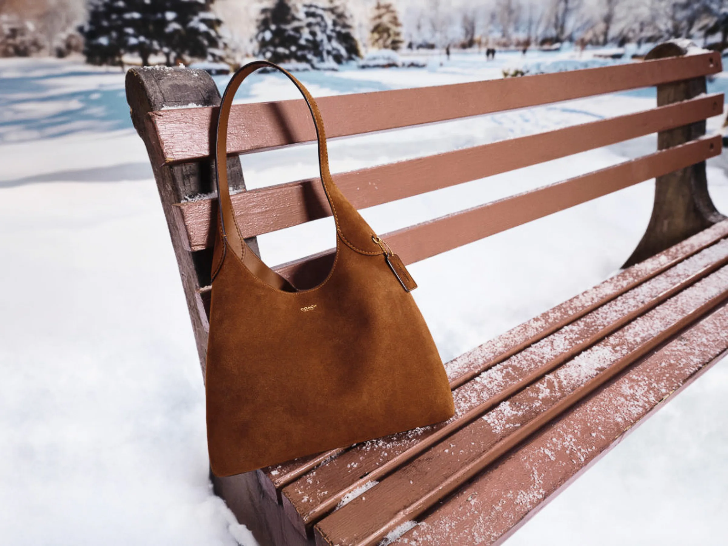 AI image of a brown suede shoulder bag hanging on a snow-dusted wooden park bench in a winter landscape, for Coach.