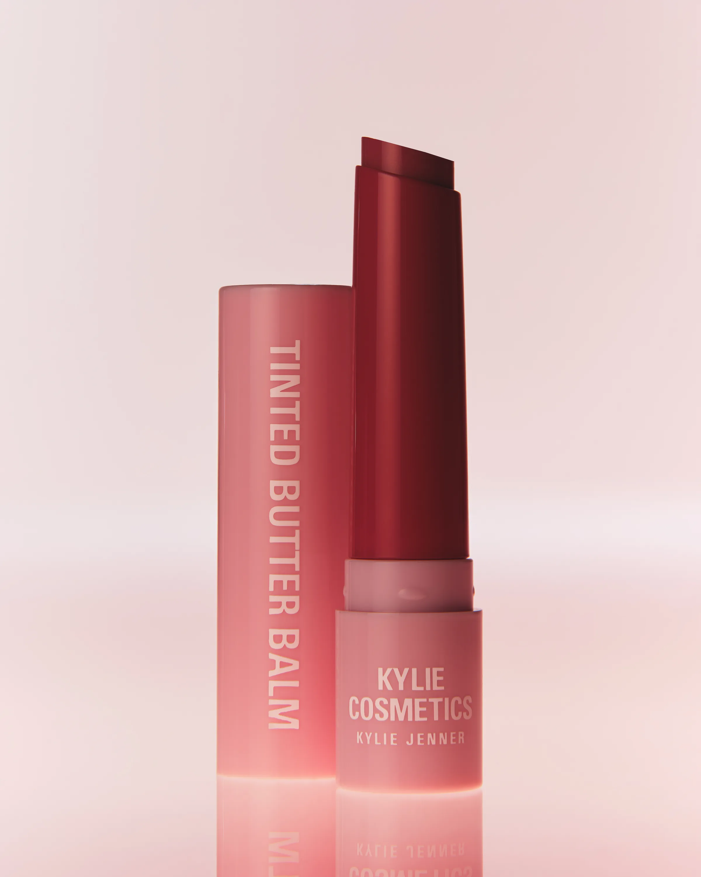 Red tinted butter balm uncapped beside its pink tube on a soft pink reflective surface, for Kylie Cosmetics.