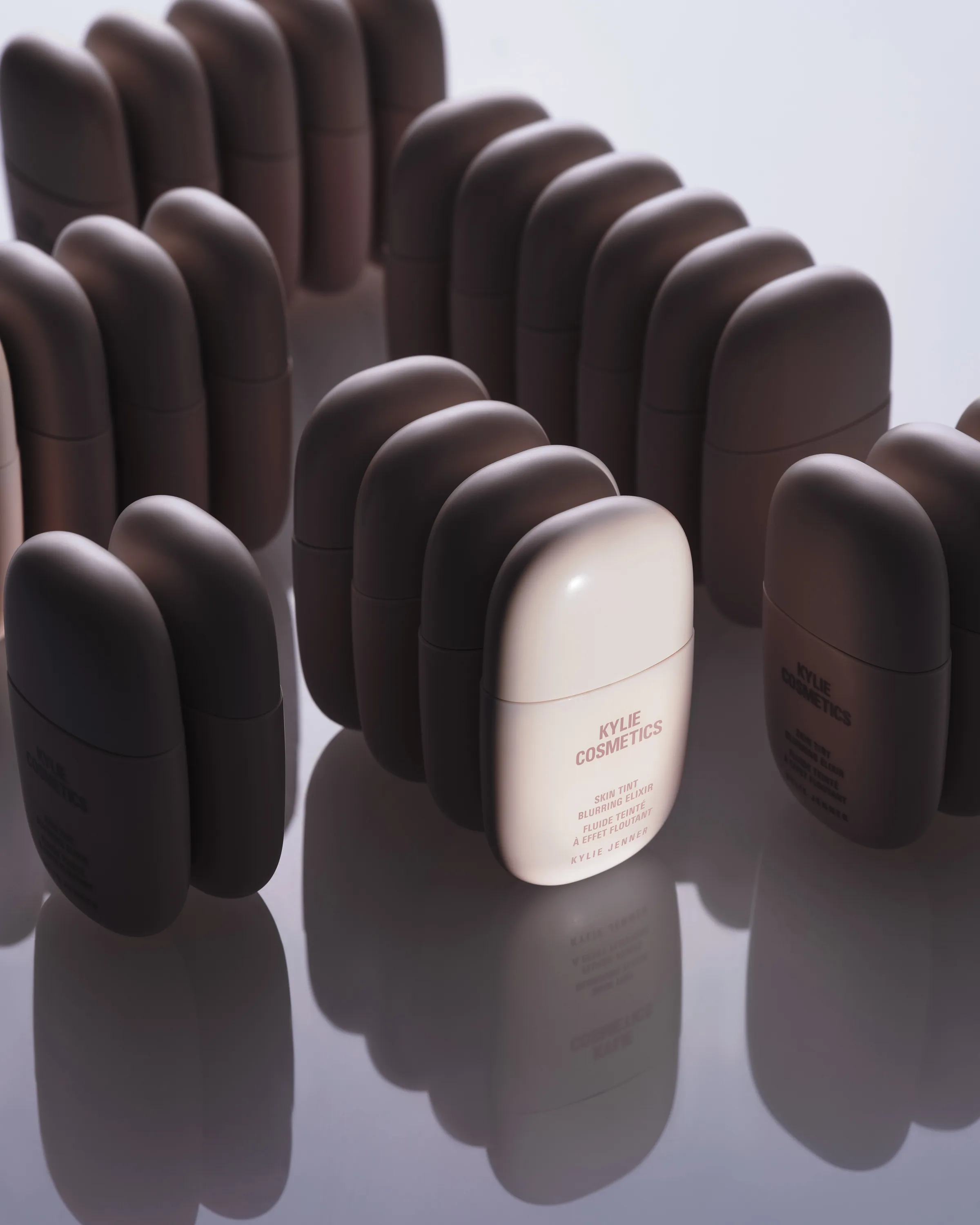 Rows of Skin Tint Blurring Elixir bottles in deep brown shades around one pale bottle on a reflective surface, for Kylie Cosmetics.