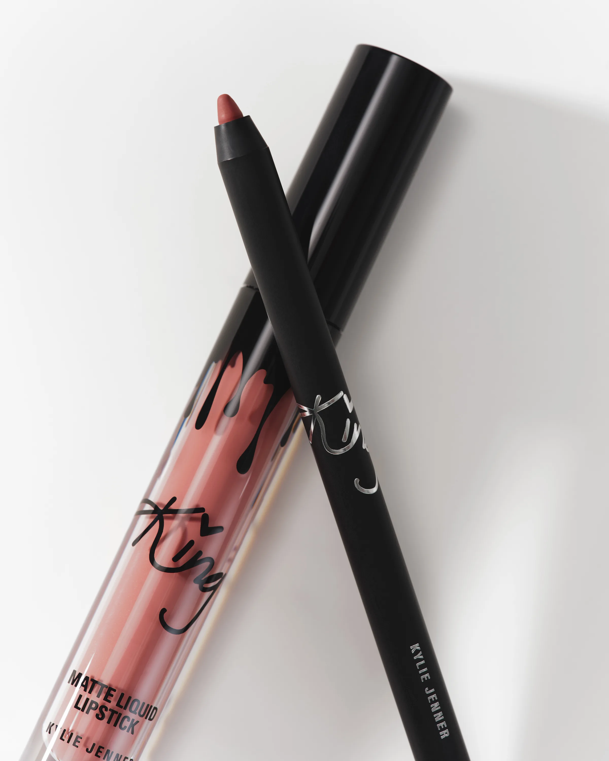 Still life of a Kylie matte liquid lipstick in a nude rose shade crossed with a black lip liner on white, for Kylie Cosmetics.