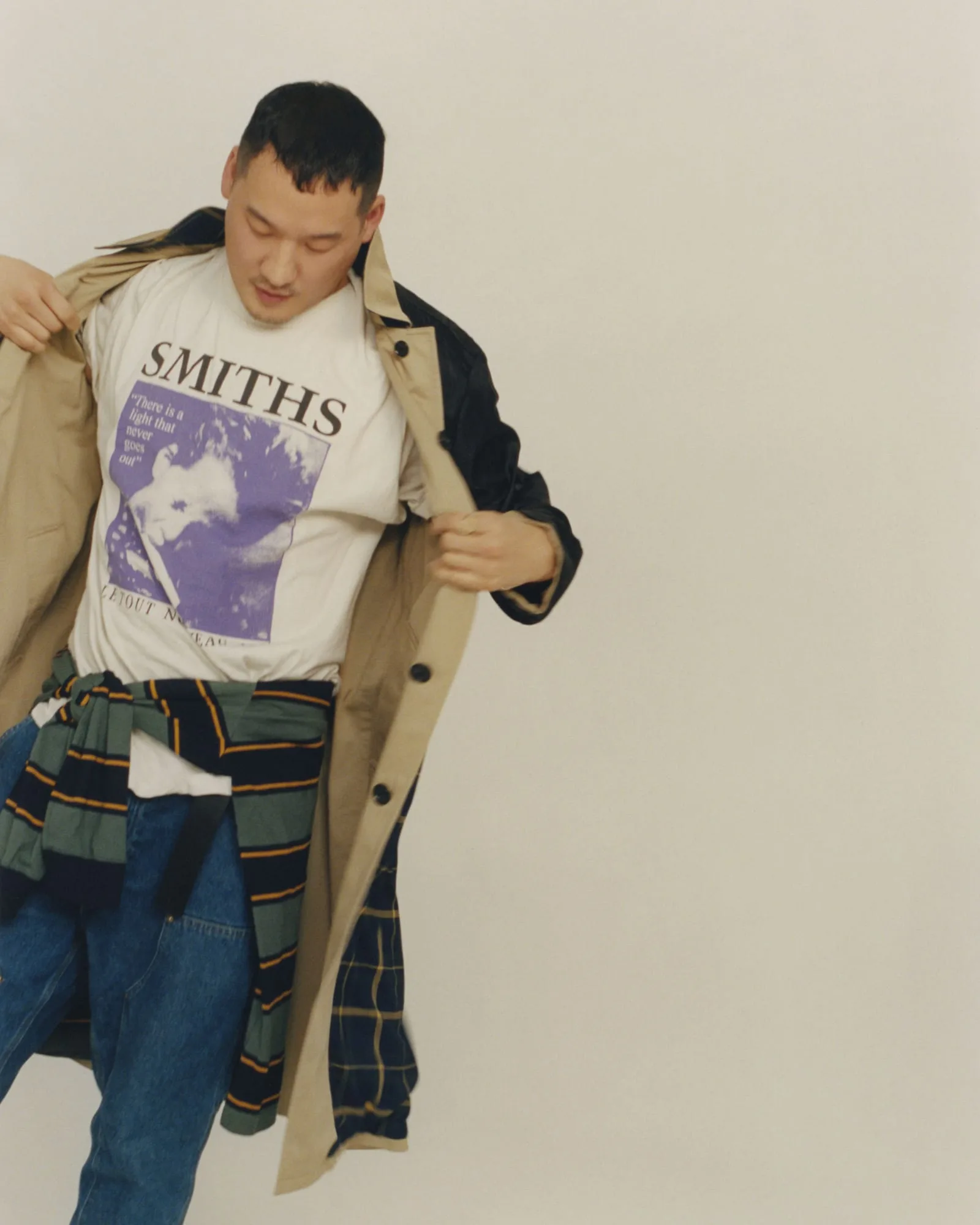 Man opening a beige trench coat over a Smiths band tee, striped sweater tied at waist, and jeans, for Vogue Korea.