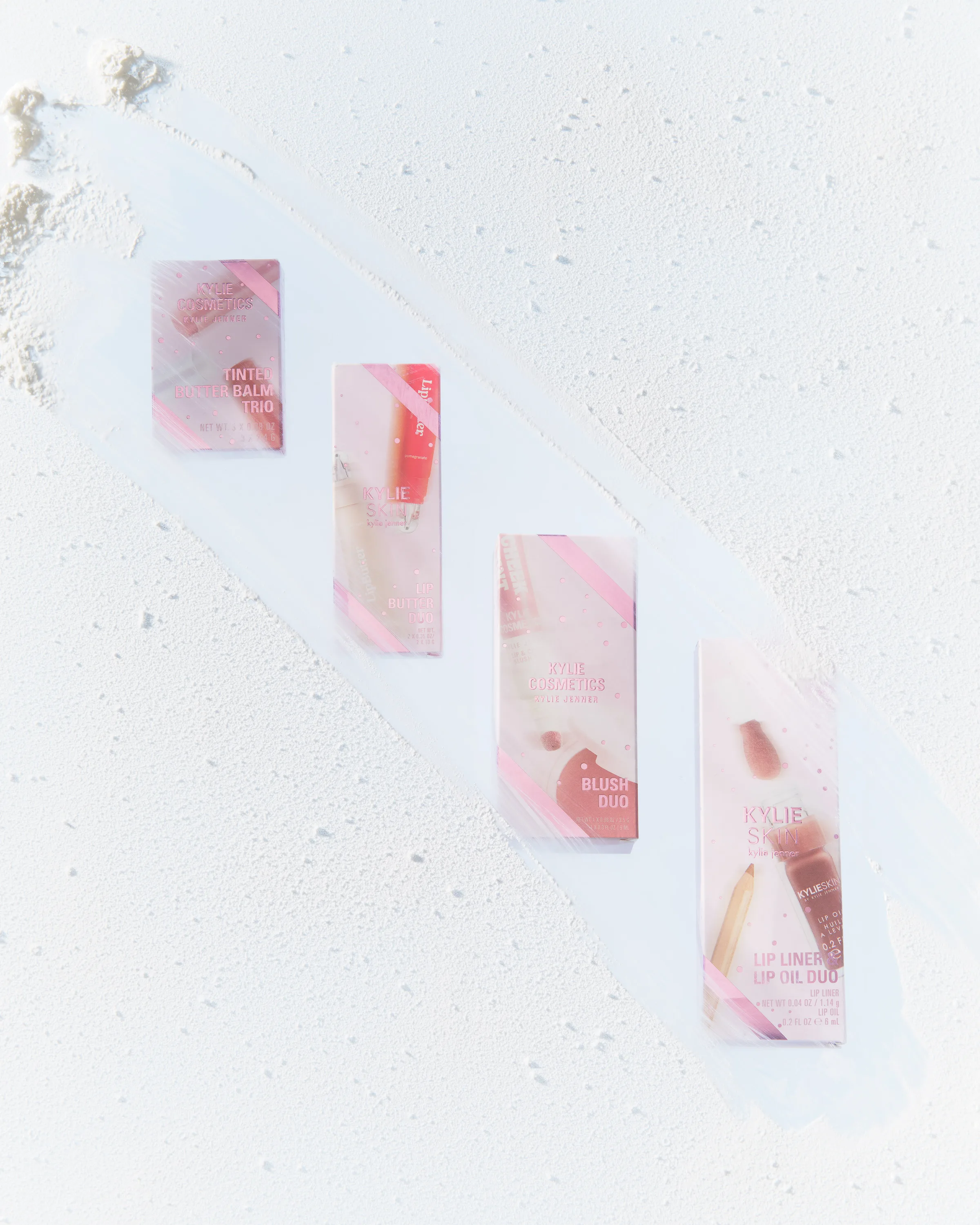 Four pink holiday makeup gift boxes laid diagonally on a frosted white surface with powdery snow, for Kylie Cosmetics.