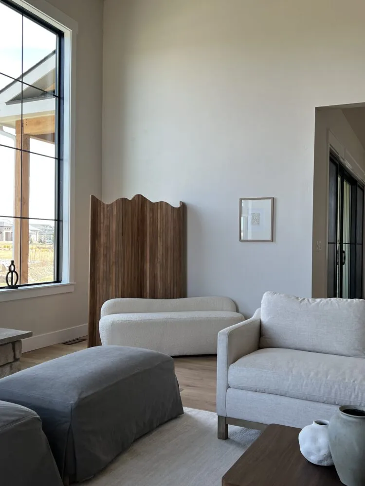 Bright living room with white armchair, boucle bench, wavy wooden folding screen and grey ottoman by a tall window.