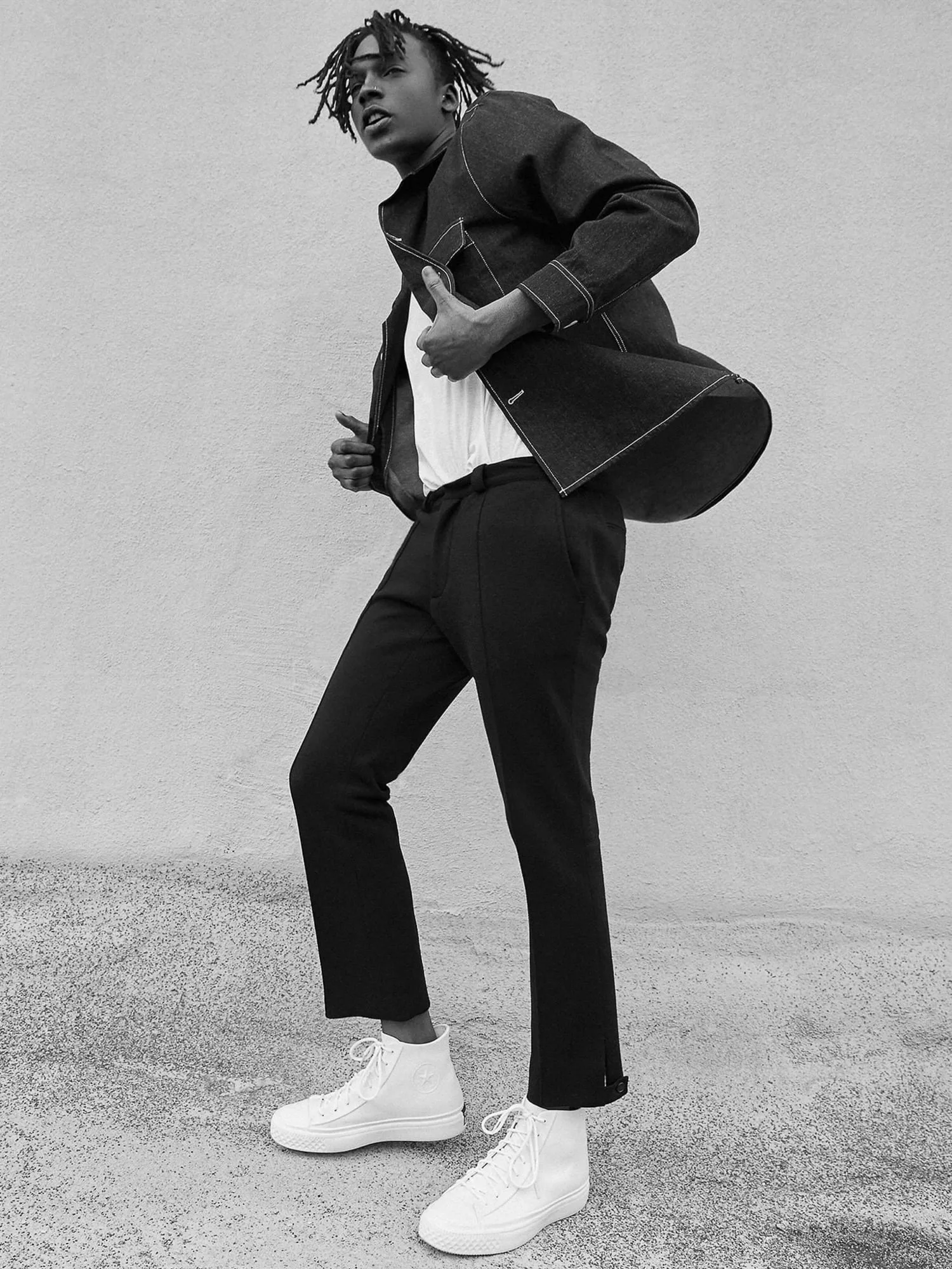 Black and white shot of a model in a dark denim jacket, black trousers and white high-top sneakers against a wall, for Converse.