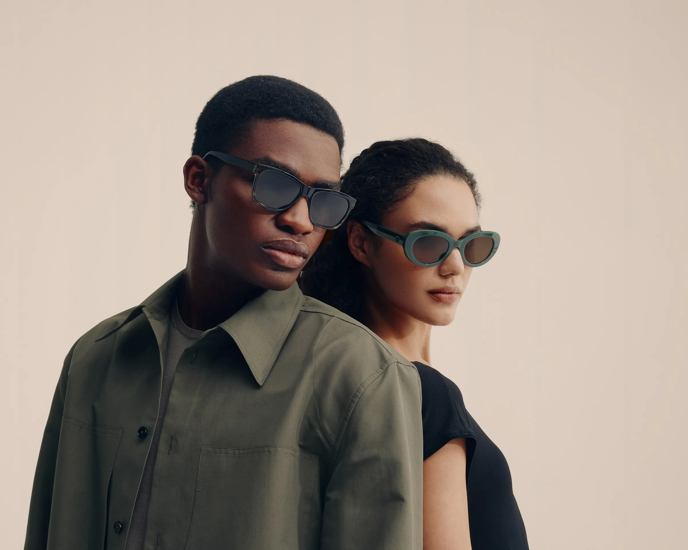 Man in olive shirt and woman in black top wearing tinted sunglasses against a beige backdrop, for Cole Haan.