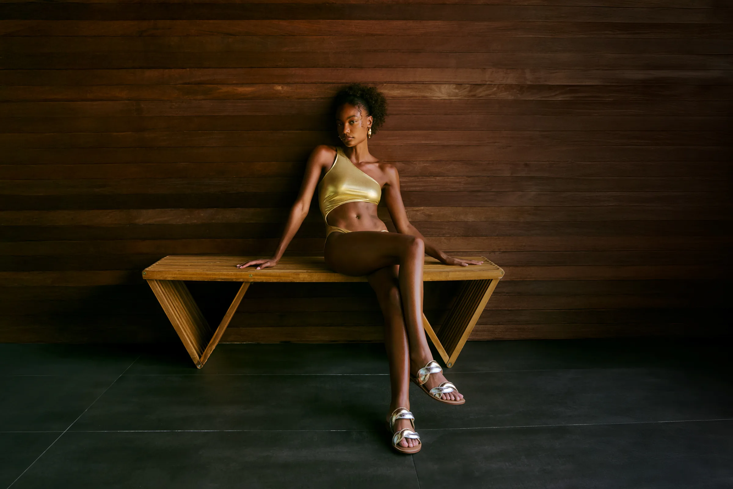 Model in a gold one-shoulder bikini and silver sandals sits on a wooden bench against a slatted wood wall, for Sam Edelman.