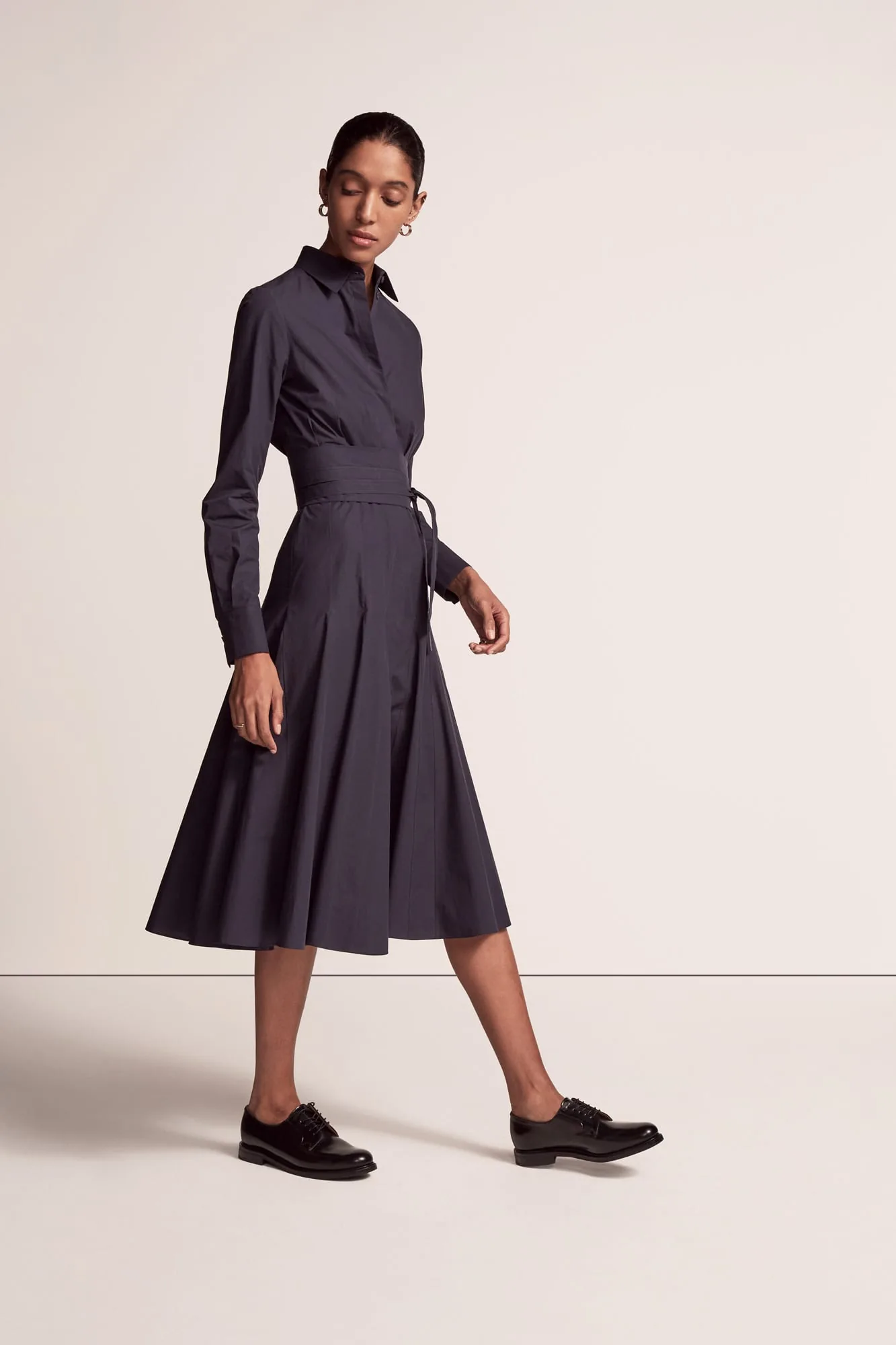 Model walking in a navy belted midi shirt dress and black leather oxfords on a cream backdrop, for Another Tomorrow.