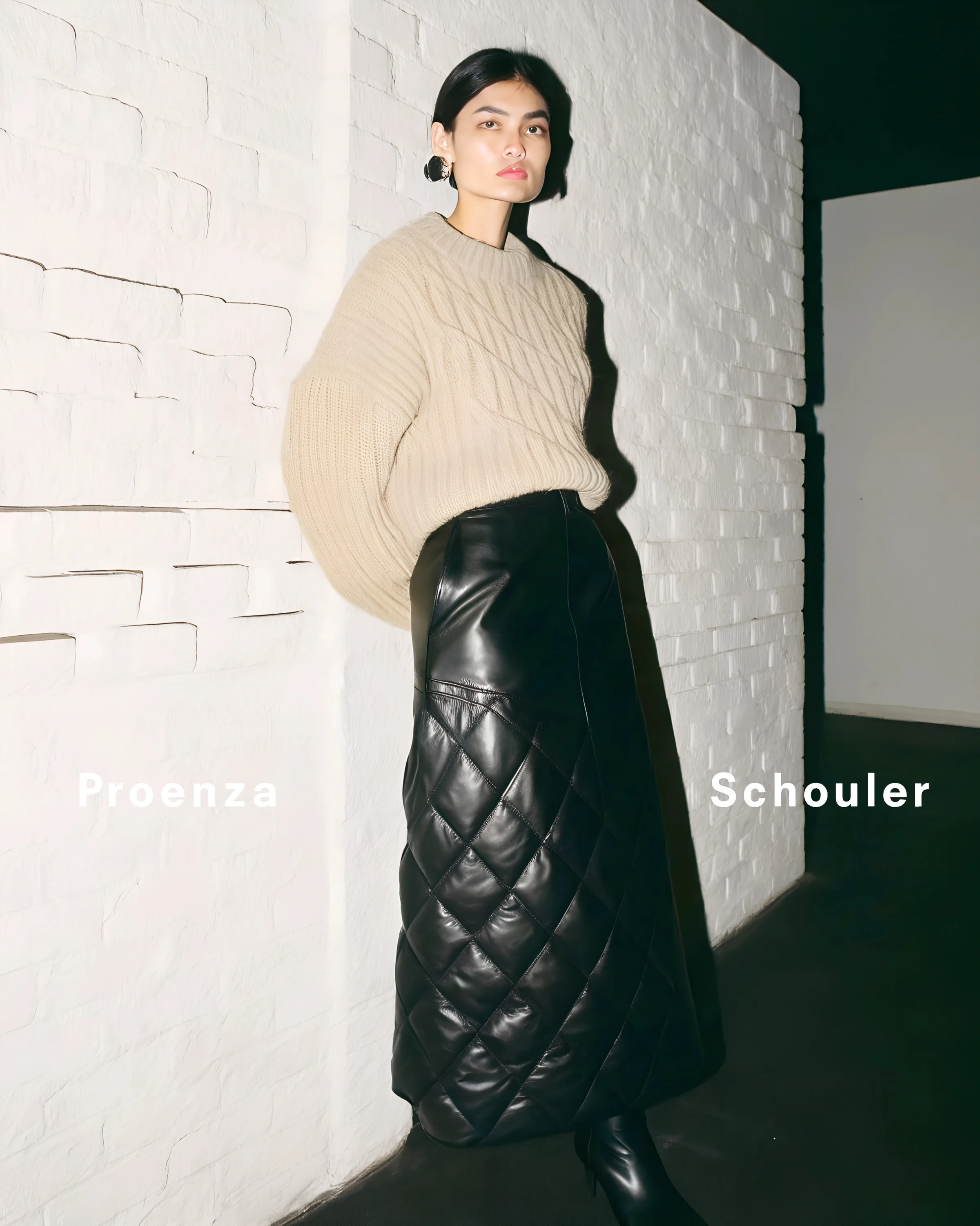 AI image of a model in a beige cable-knit sweater and black quilted leather skirt against a white brick wall. Proenza Schouler AI Concept.