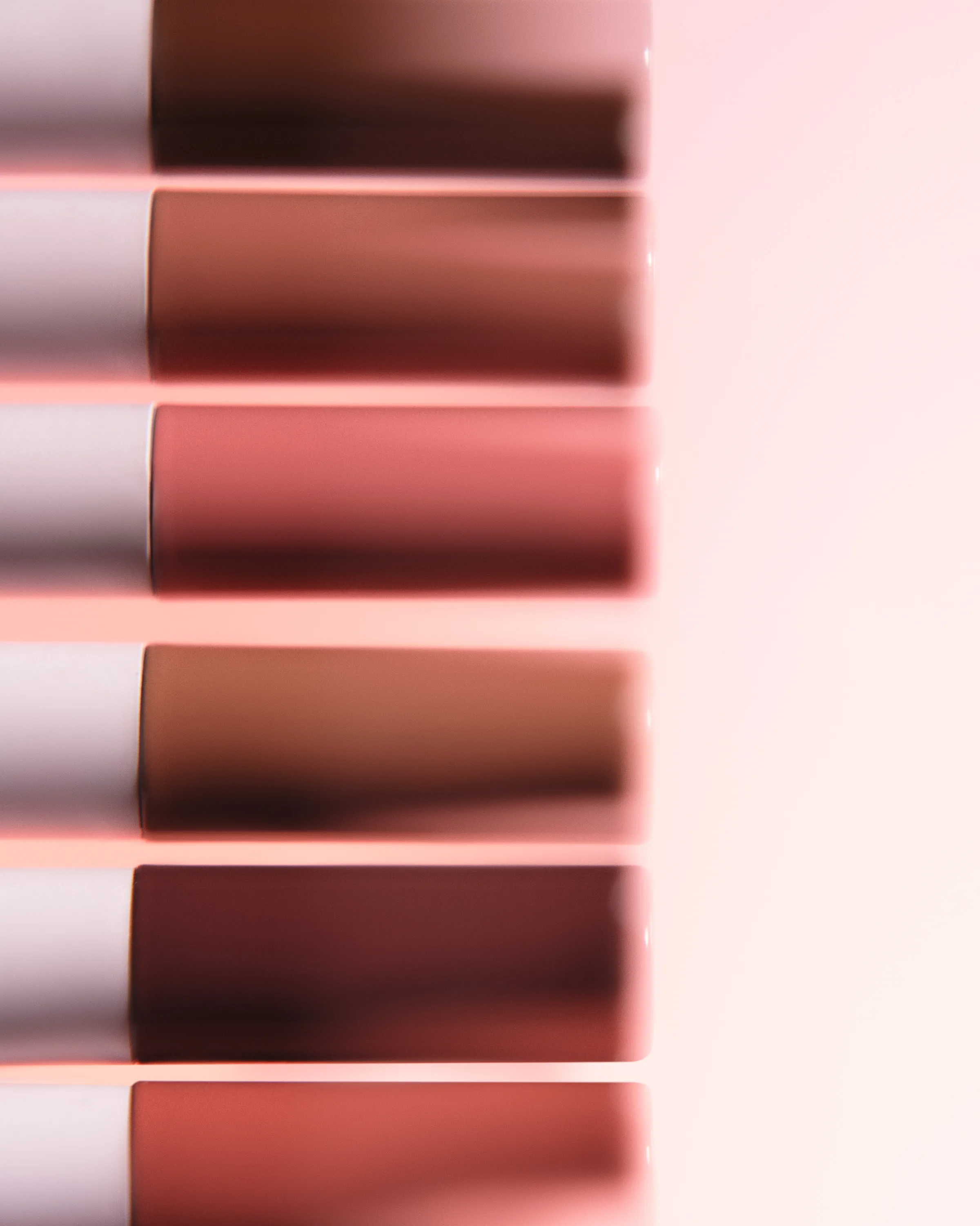 Six lip liners in nude, rose and brown shades lined up in soft focus against a pale pink backdrop, for Kylie Cosmetics.
