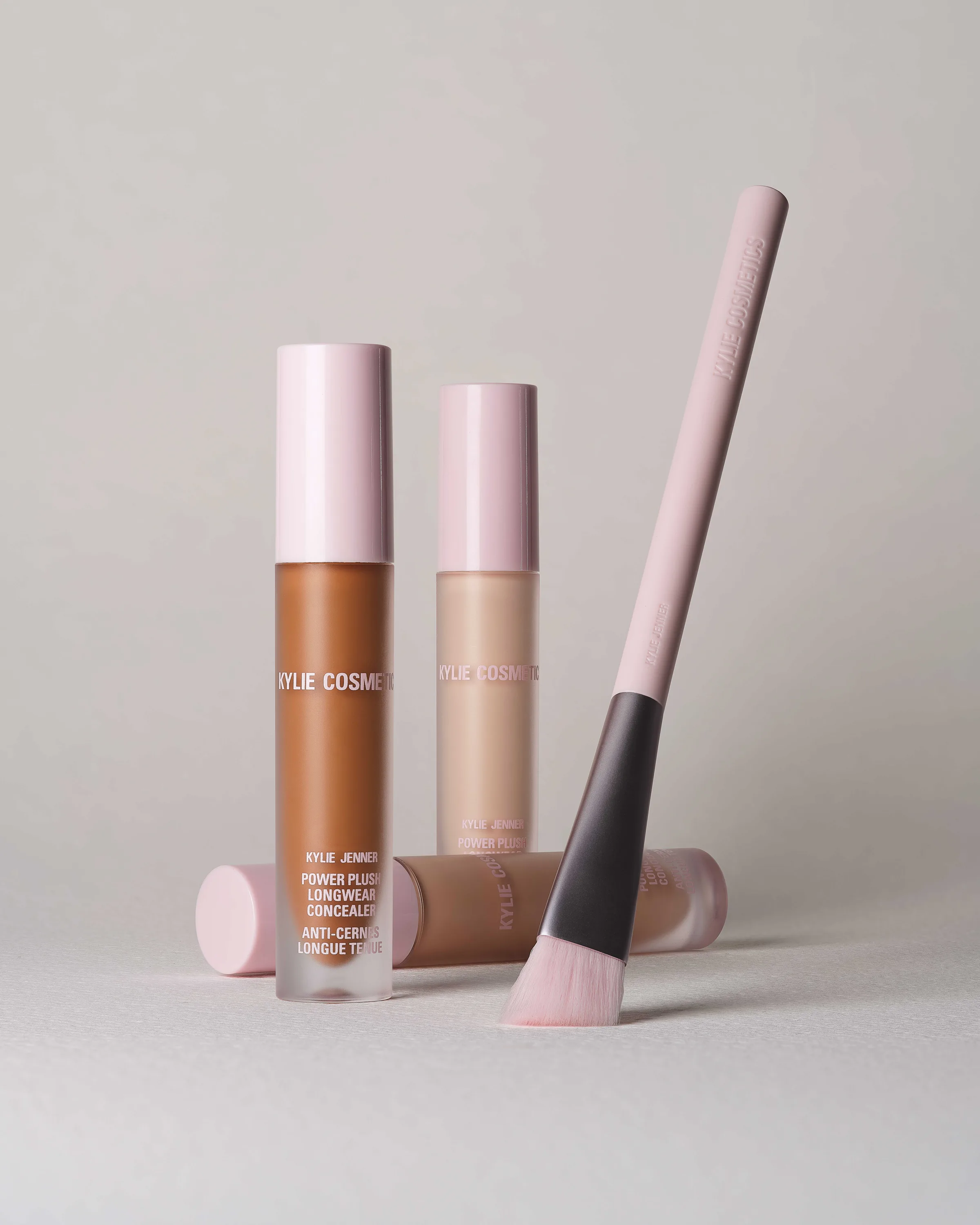 Still life of Power Plush concealer tubes in three shades with a pink-handled brush on grey, for Kylie Cosmetics.