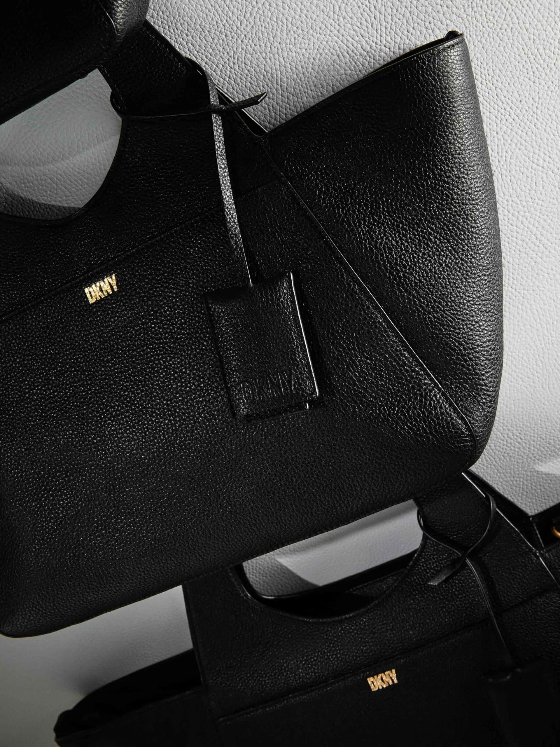 Black pebbled leather tote bags with gold logo and hanging leather tag, stacked against a grey wall, for DKNY.